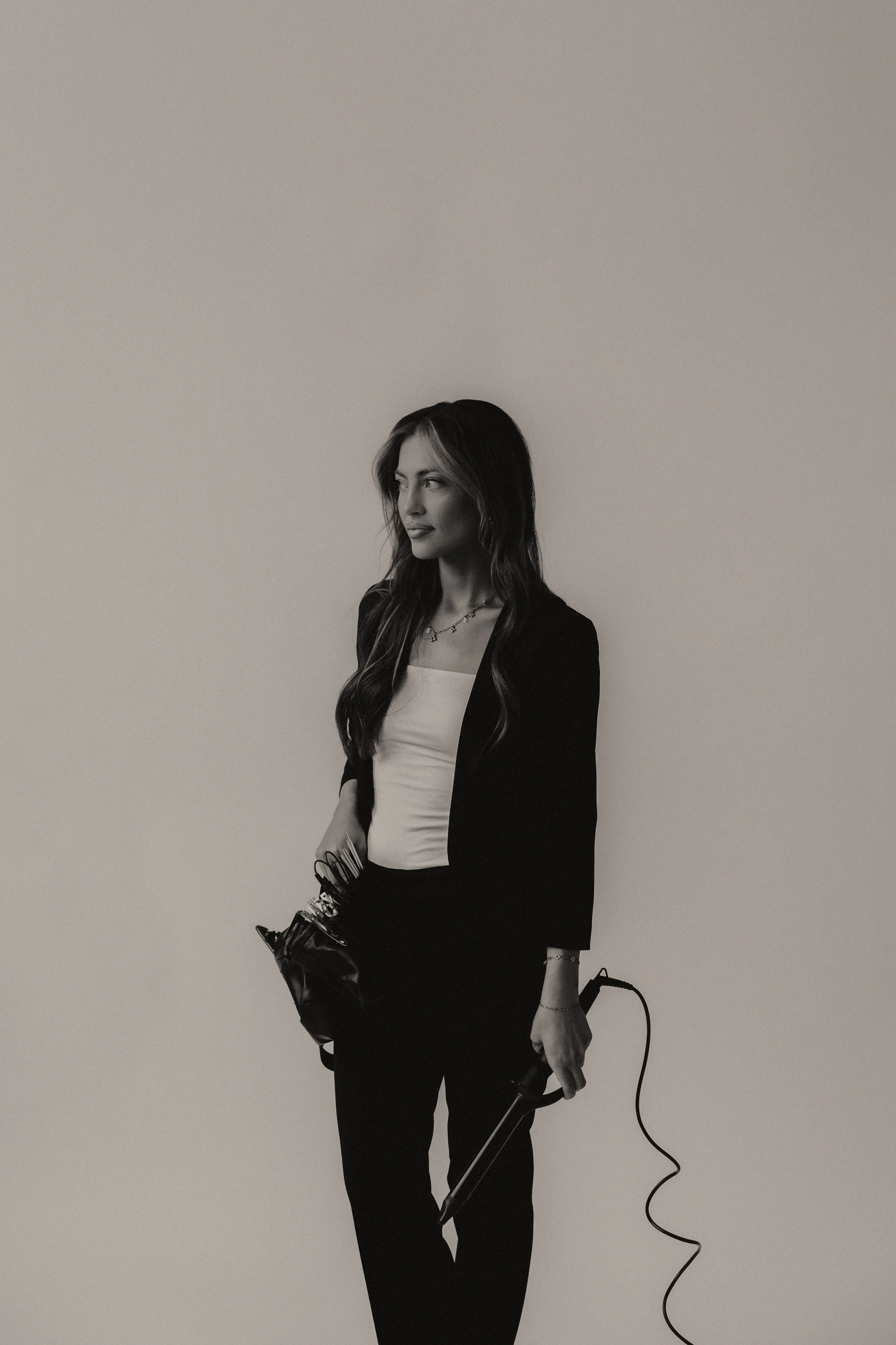 Black-and-white photo of a woman with long hair, wearing a blazer and trousers, holding a vacuum cleaner with the cord hanging down, standing against a plain background.