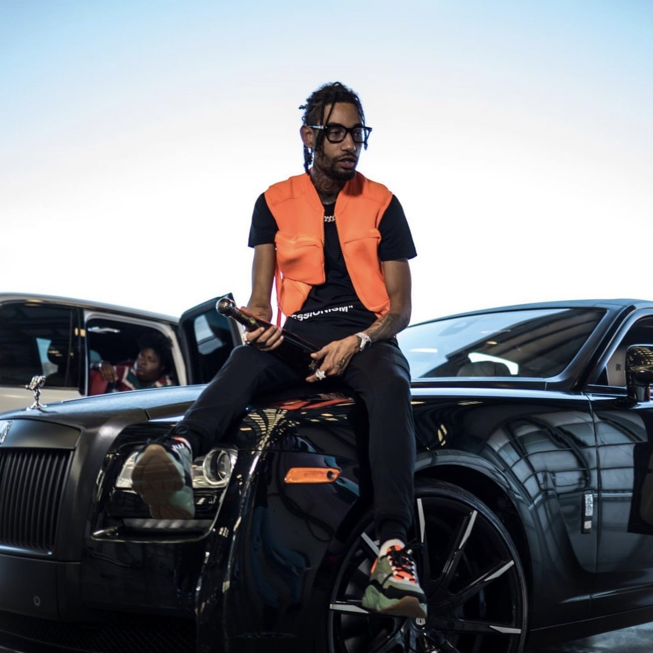 Man with dreadlocks wearing glasses and an orange vest sitting on a black luxury car, holding a trophy, with another car and a person inside in the background.