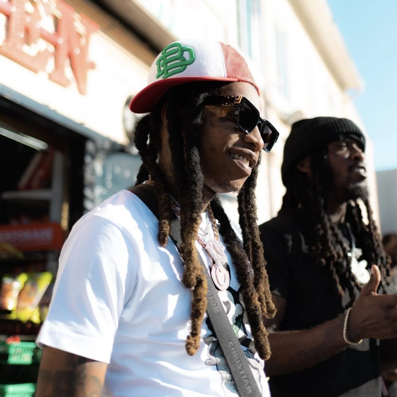 Two men with dreadlocks, sunglasses, and jewelry standing outdoors with a building and bright sky in the background, one wearing a red and white cap and a white t-shirt.