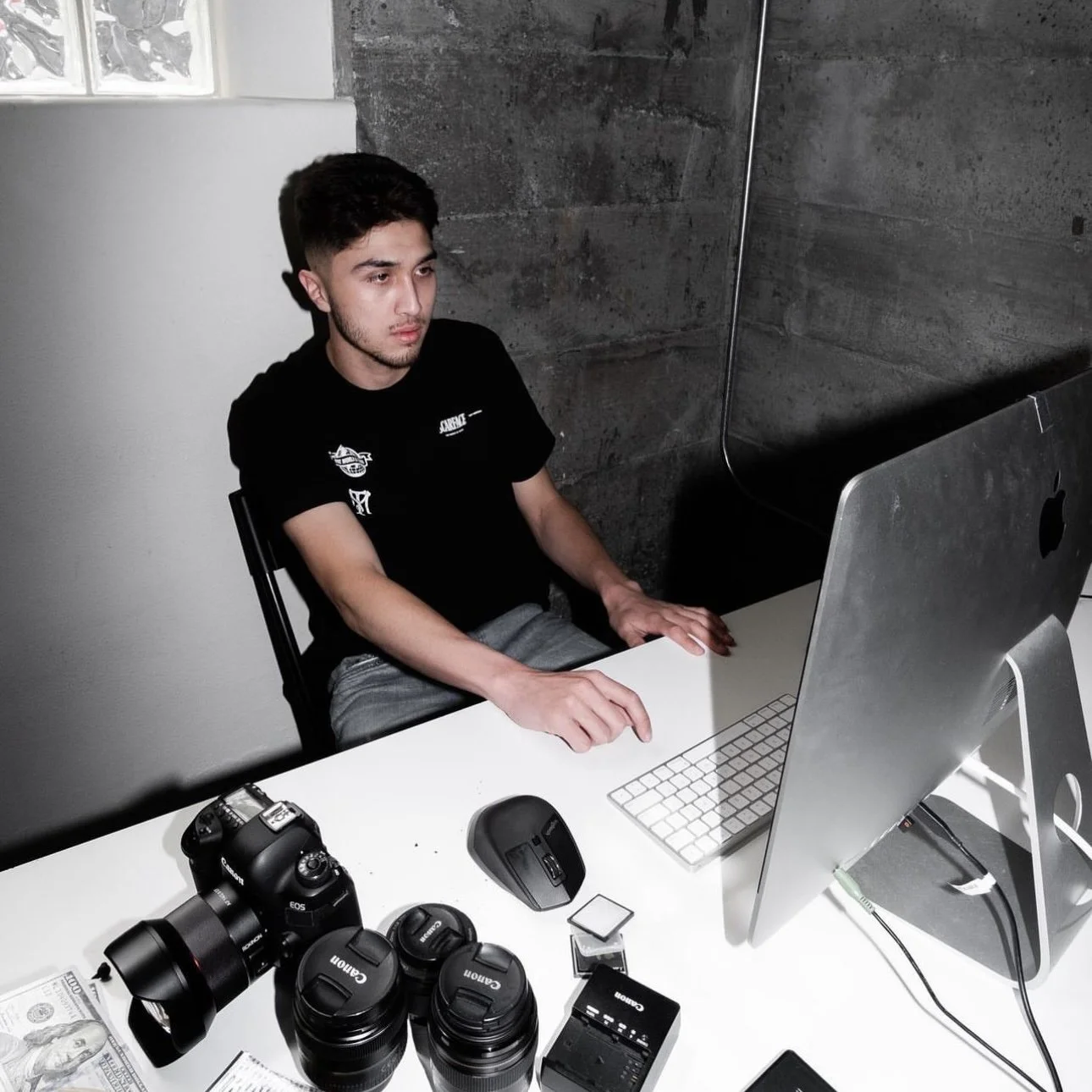 Teenage boy sitting at a white desk with a computer, surrounded by cameras, camera lenses, and other photography equipment.