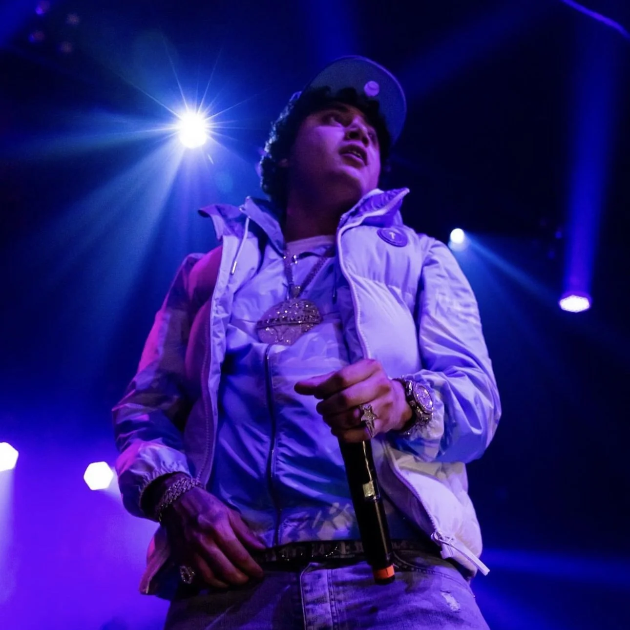 A male rapper performing on stage illuminated by blue and purple stage lights, wearing a white jacket, a baseball cap, large jewelry, and holding a microphone.