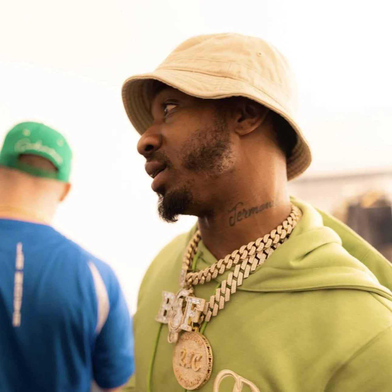 A man wearing a beige bucket hat, lime green hoodie, and several gold chains with large pendants, including a medallion with 
