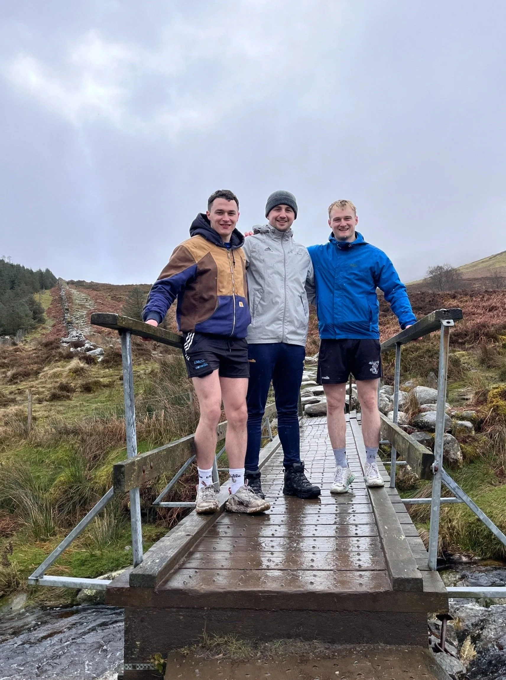 Three people standing on a small wooden bridge outdoors with a grassy hill and cloudy sky in the background.
