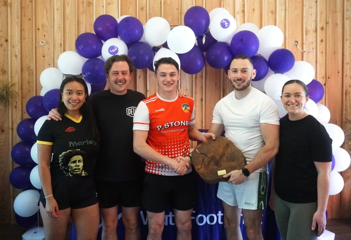 Group of five people standing in front of a background of purple and white balloons, with one person holding a wooden plaque.