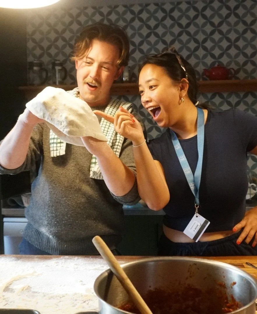 Two people in a kitchen, holding and examining a piece of dough, smiling, with a pot on the counter.