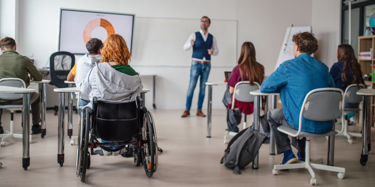 A teacher stands explaining to his class in front of a whiteboard with a pie chart. Students, including one in a wheelchair, attentively face the teacher.