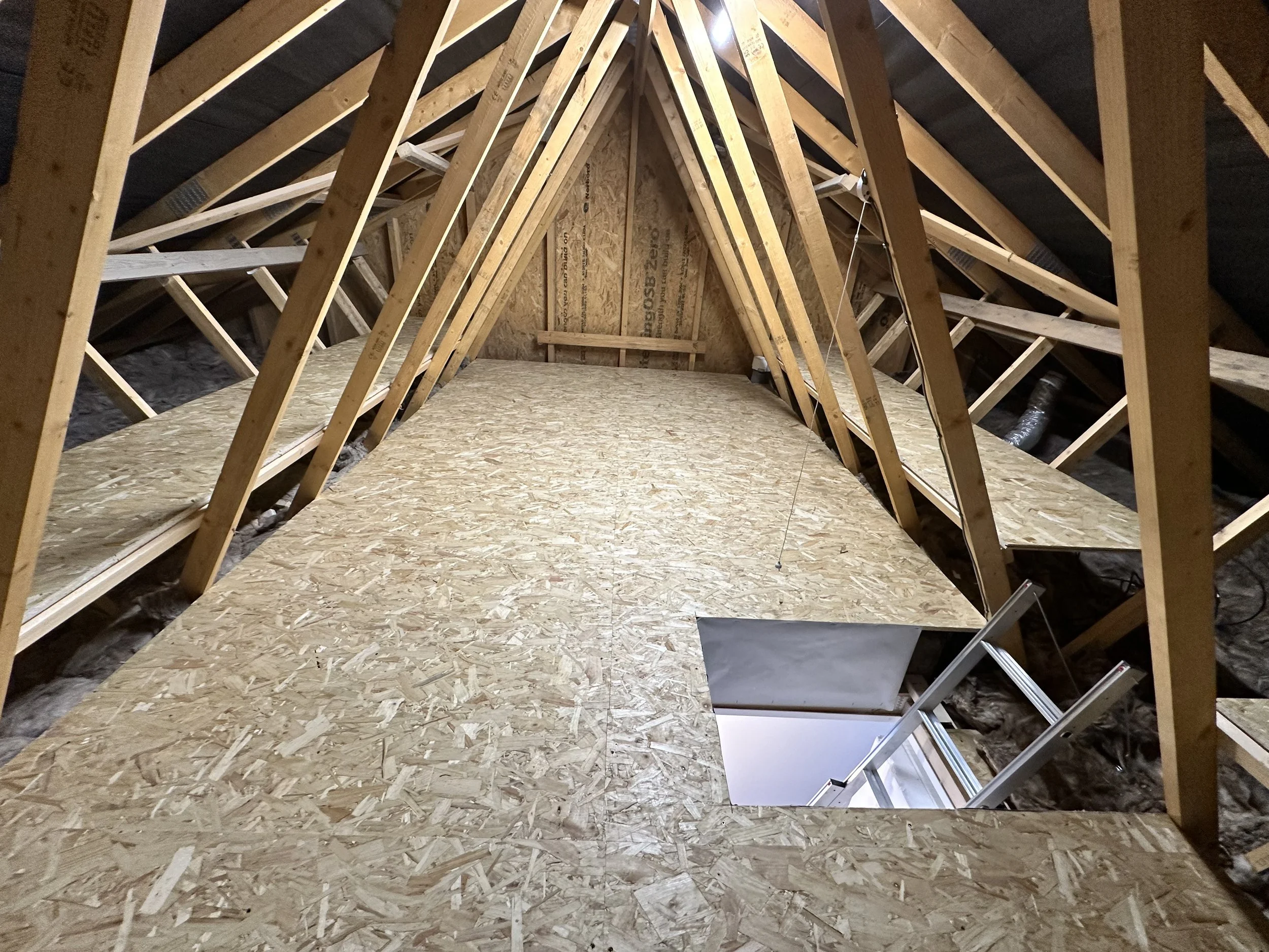 Attic with wooden rafters and plywood flooring, includes a small window with a ladder leaning against the floor.
