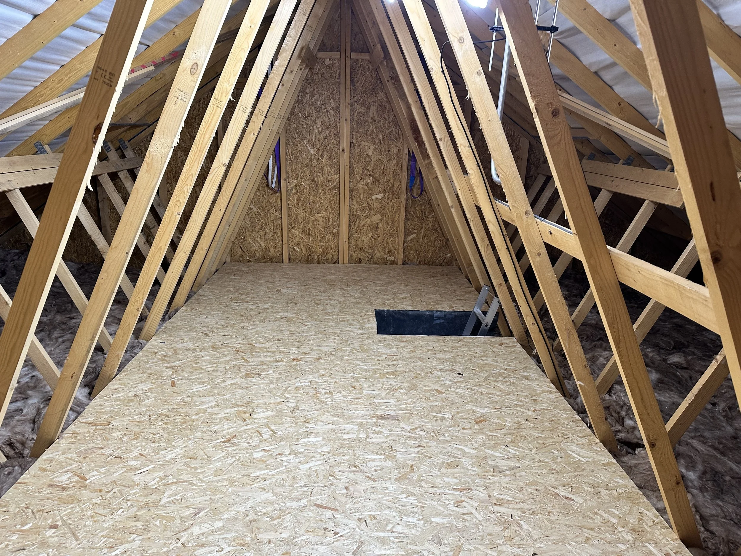 The interior of an attic under construction with wooden framing, OSB flooring, insulation around the edges, and a small foldable step ladder.