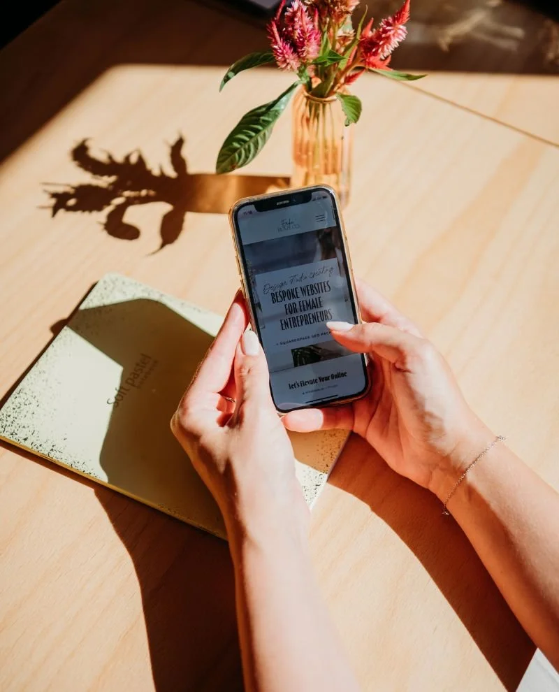 Person holding a smartphone over a notebook on a wooden table. A vase with pink and red flowers casts a shadow on the table.