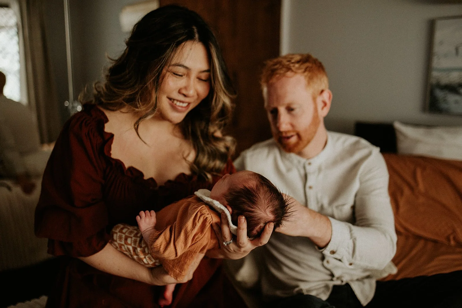 A mother and father hold their newborn baby whilst being photographed in their Brisbane home. They are relaxed and the image is of a lifestyle nature with beautiful colours and styling.