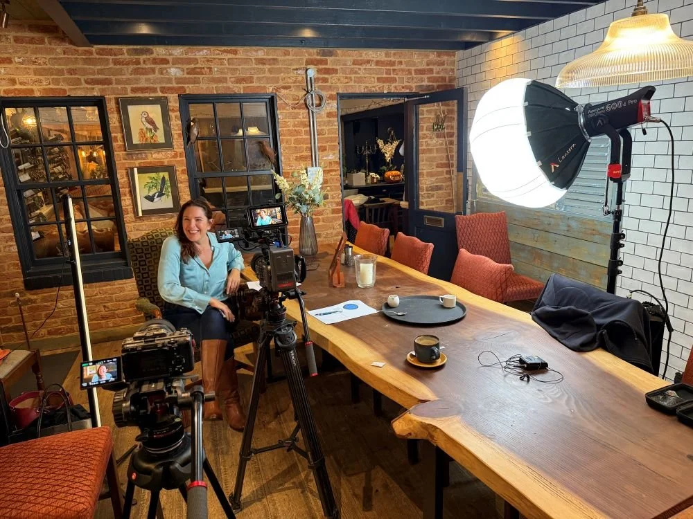 A lady sat at a wooden table with video cameras and lighting around her for a video shoot.