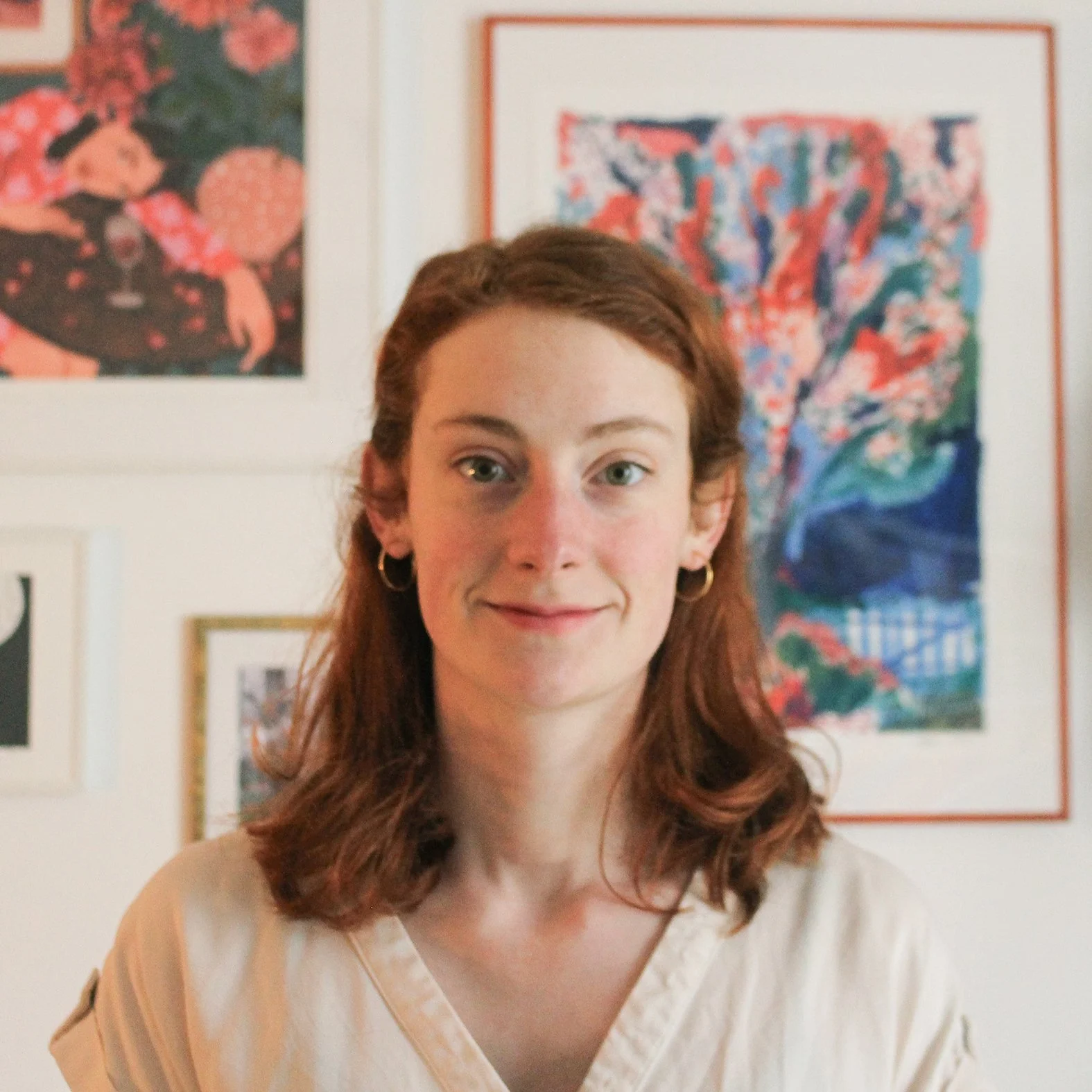 Photographie de Mathilde Villerot, illustratrice et sophrologue à Nantes. Jeune femme aux cheveux roux souriante, portant un chemisier blanc, avec des œuvres d'art colorées en arrière-plan.