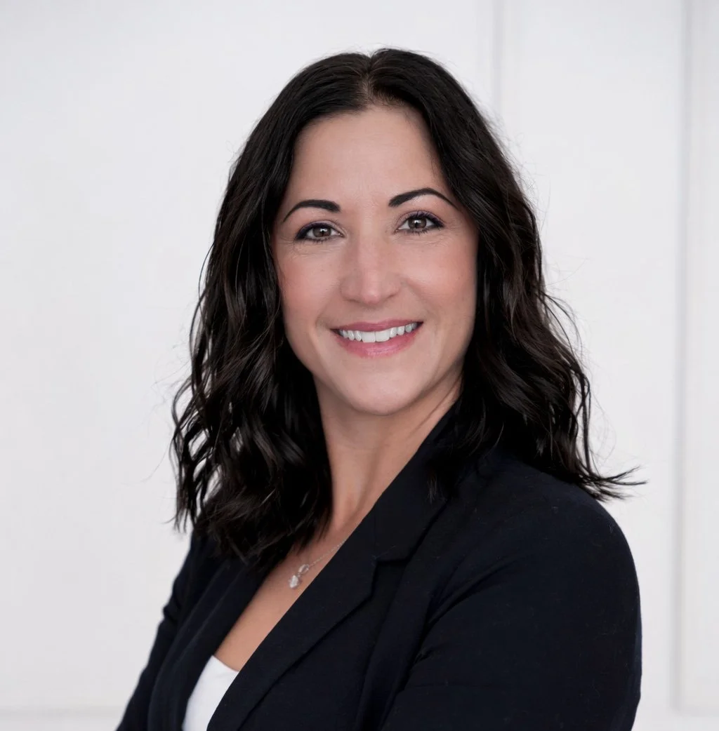 A woman with shoulder-length dark wavy hair smiling at the camera, wearing a black blazer and a white top, against a plain light background.