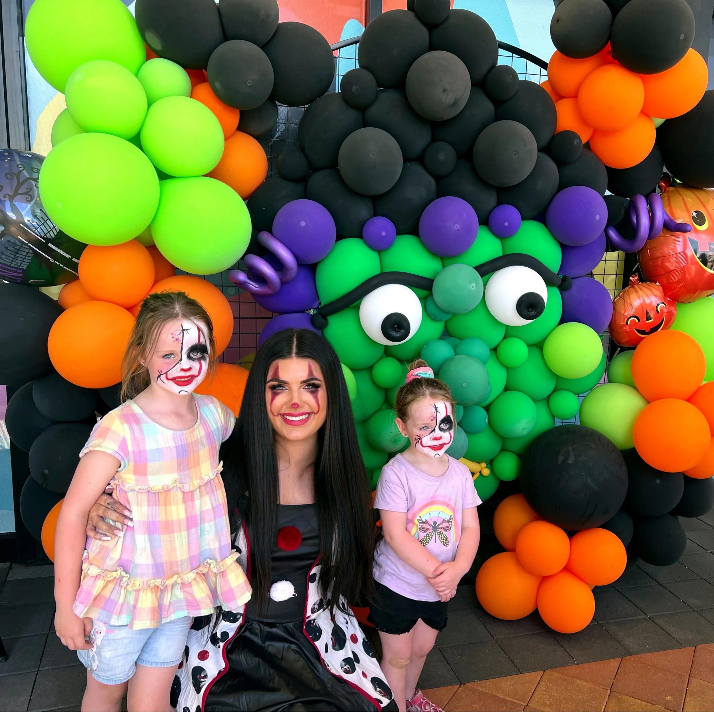 Woman and two girls in Halloween costumes posing in front of a colorful balloon display, with face paint and dressed as spooky characters.