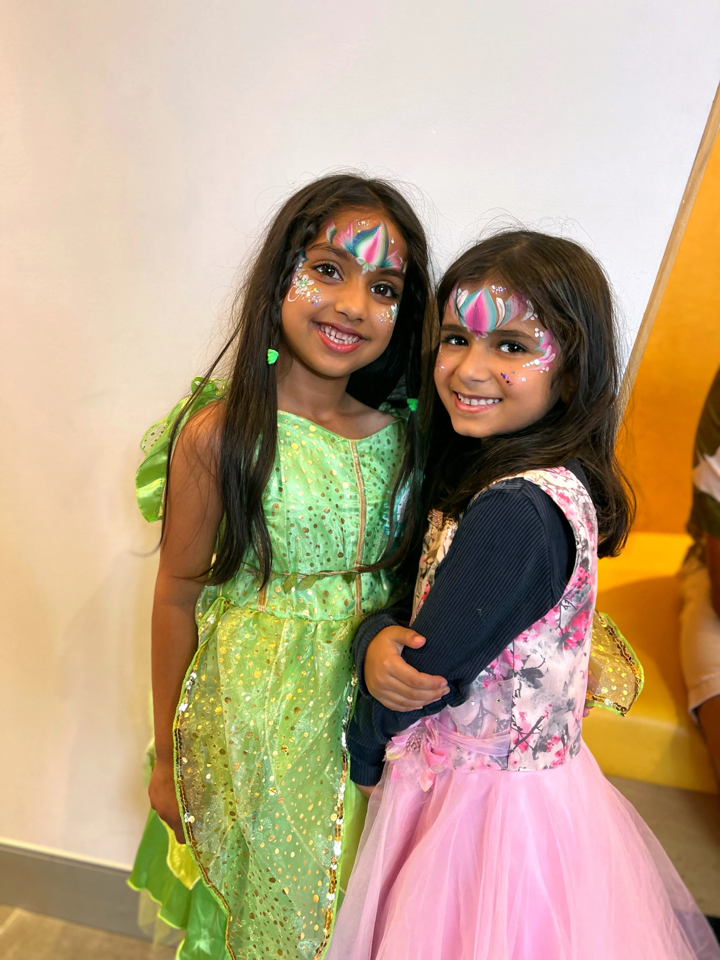 Two young girls with face paint and colorful dresses smiling indoors.