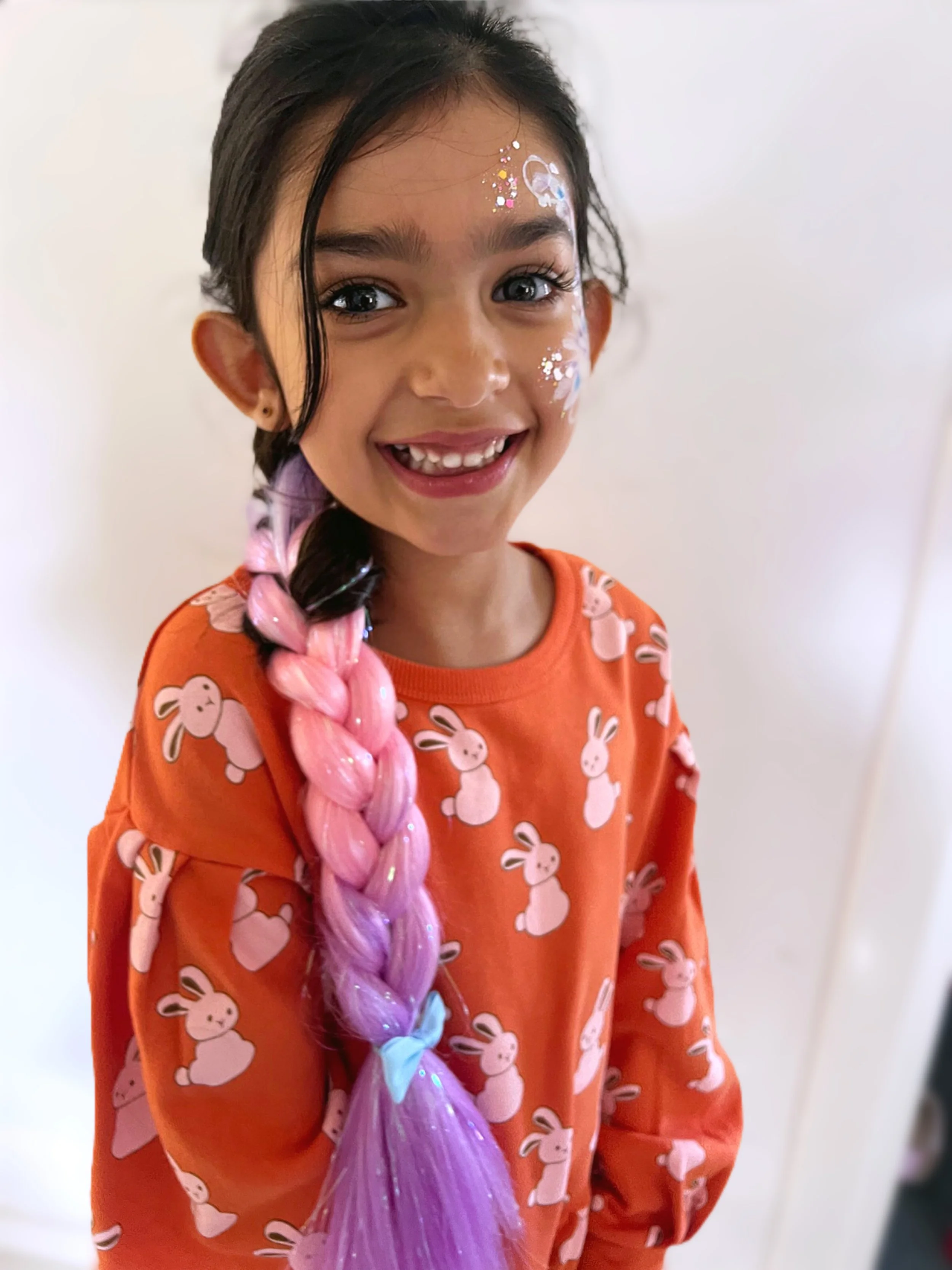 Smiling young girl with pink and purple braided hair, wearing an orange shirt with bunny print, with face paint and glitter, standing against a plain white background.