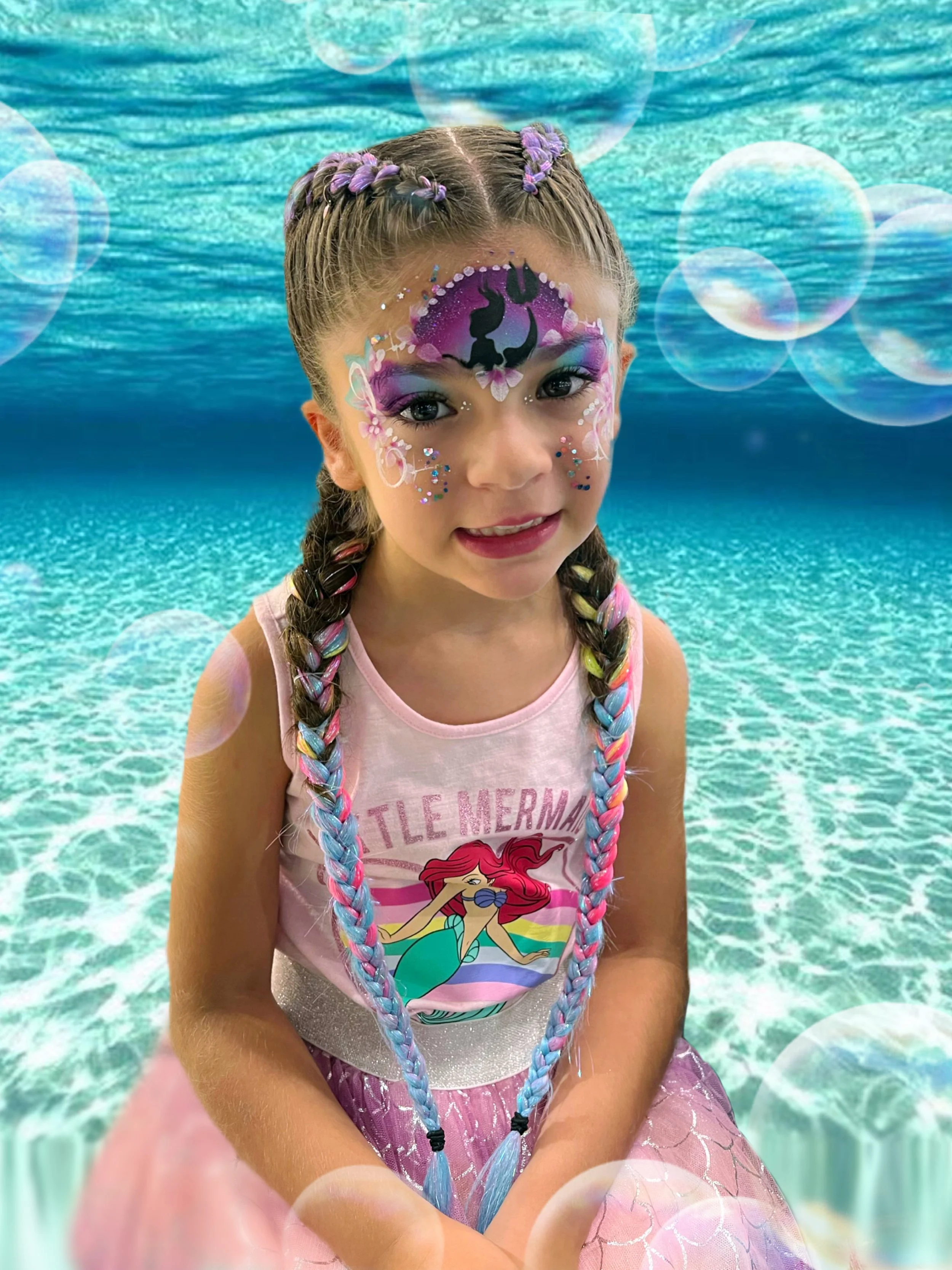 A young girl with colorful face paint and braided hair sitting in front of a digitally added ocean background with bubbles.