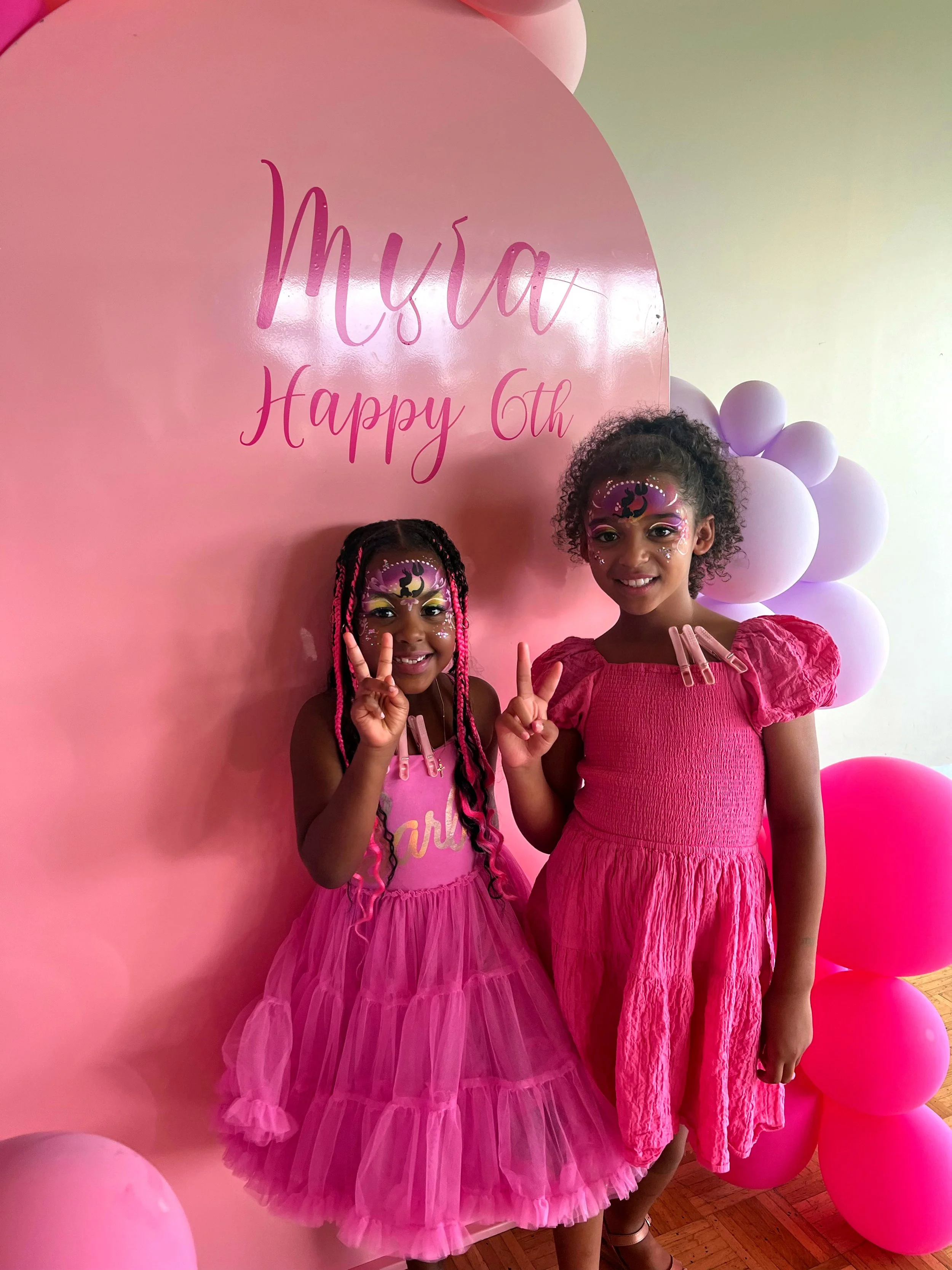 Two young girls dressed in pink with face paint, standing in front of a pink backdrop with balloons, celebrating a sixth birthday.