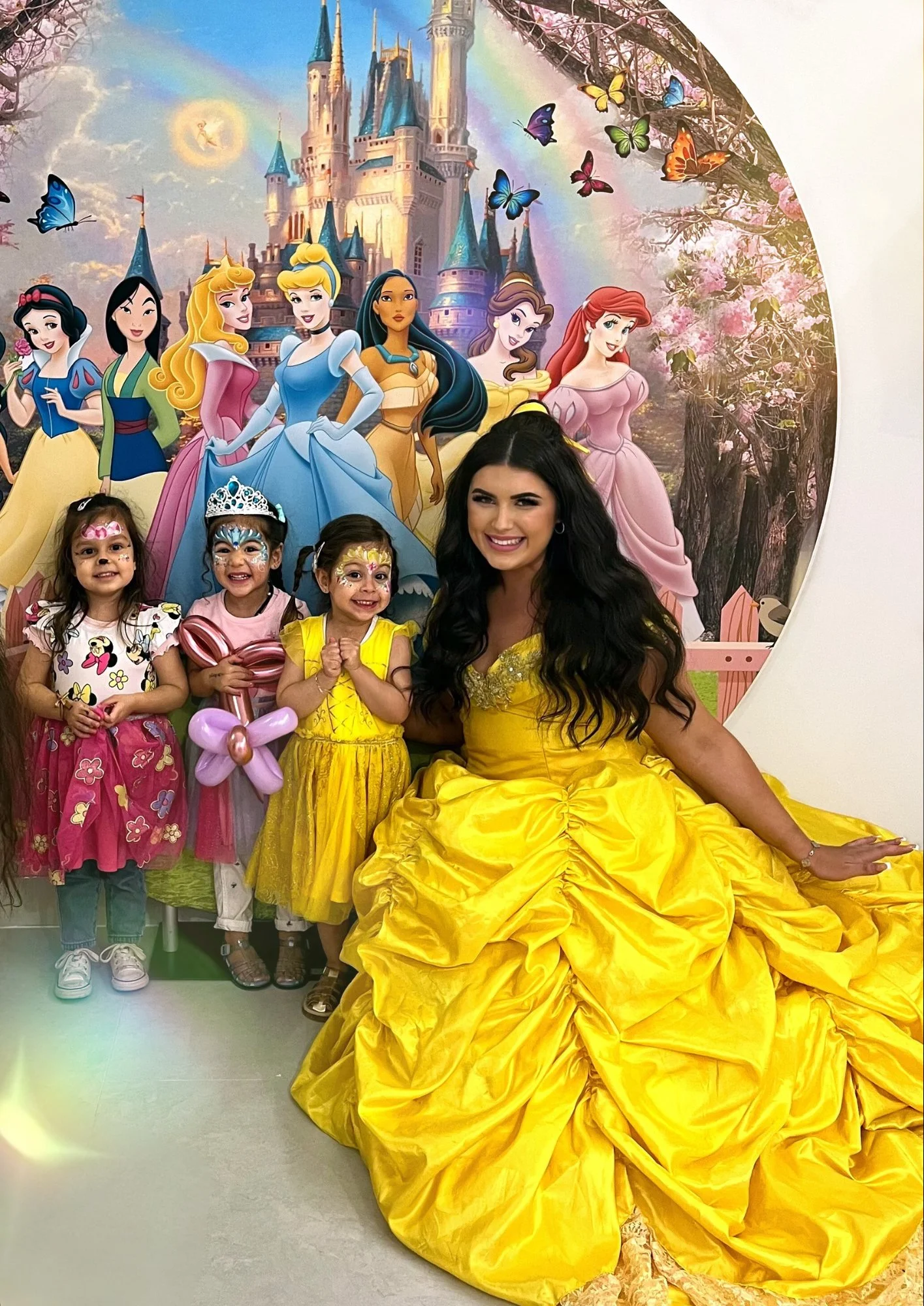 A woman dressed as Belle from Disney's Beauty and the Beast sitting beside three young girls with face paint and Princess-themed outfits, at a themed party with a Disney princess mural in the background.