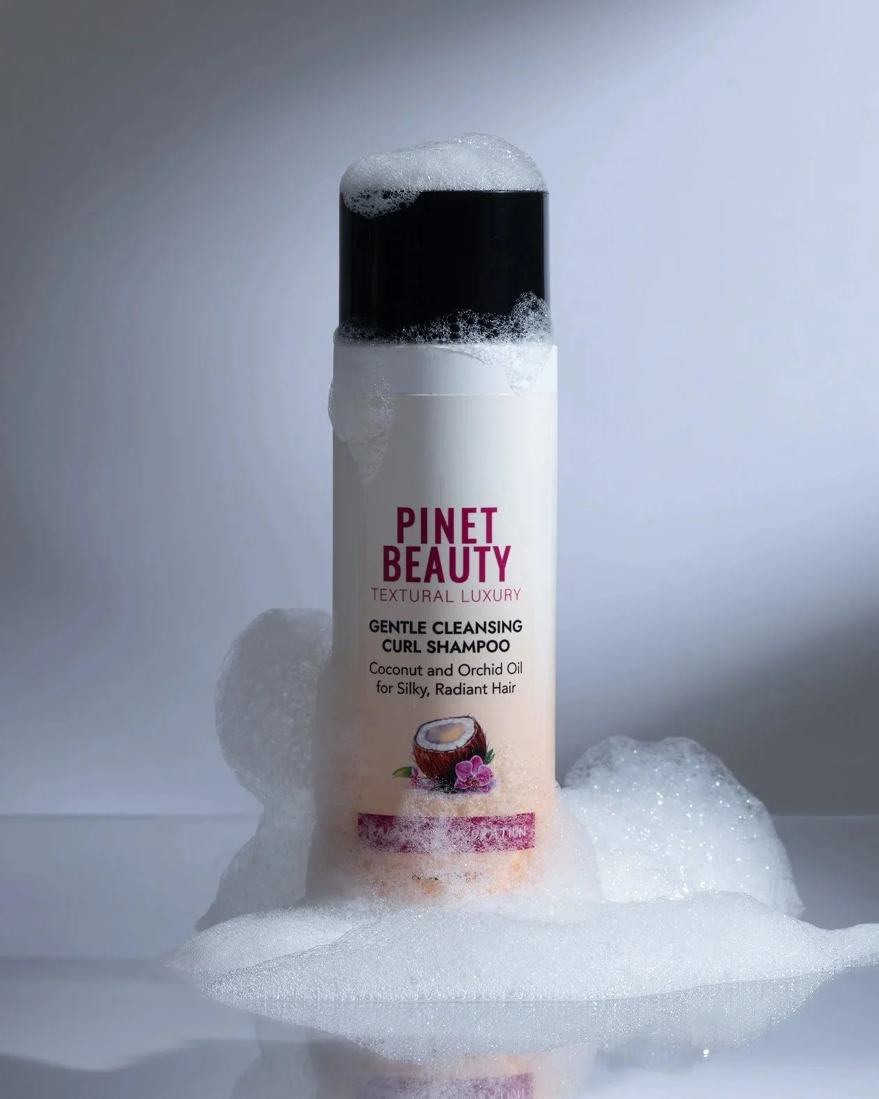 A bottle of Pinet Beauty gentle cleansing curl shampoo surrounded by soap suds on a reflective surface. product photography portfolio. 