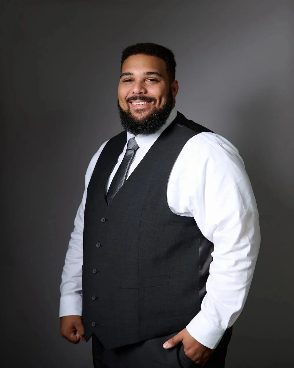 Portrait of a smiling man in a white shirt, black vest, and tie against a gray background.