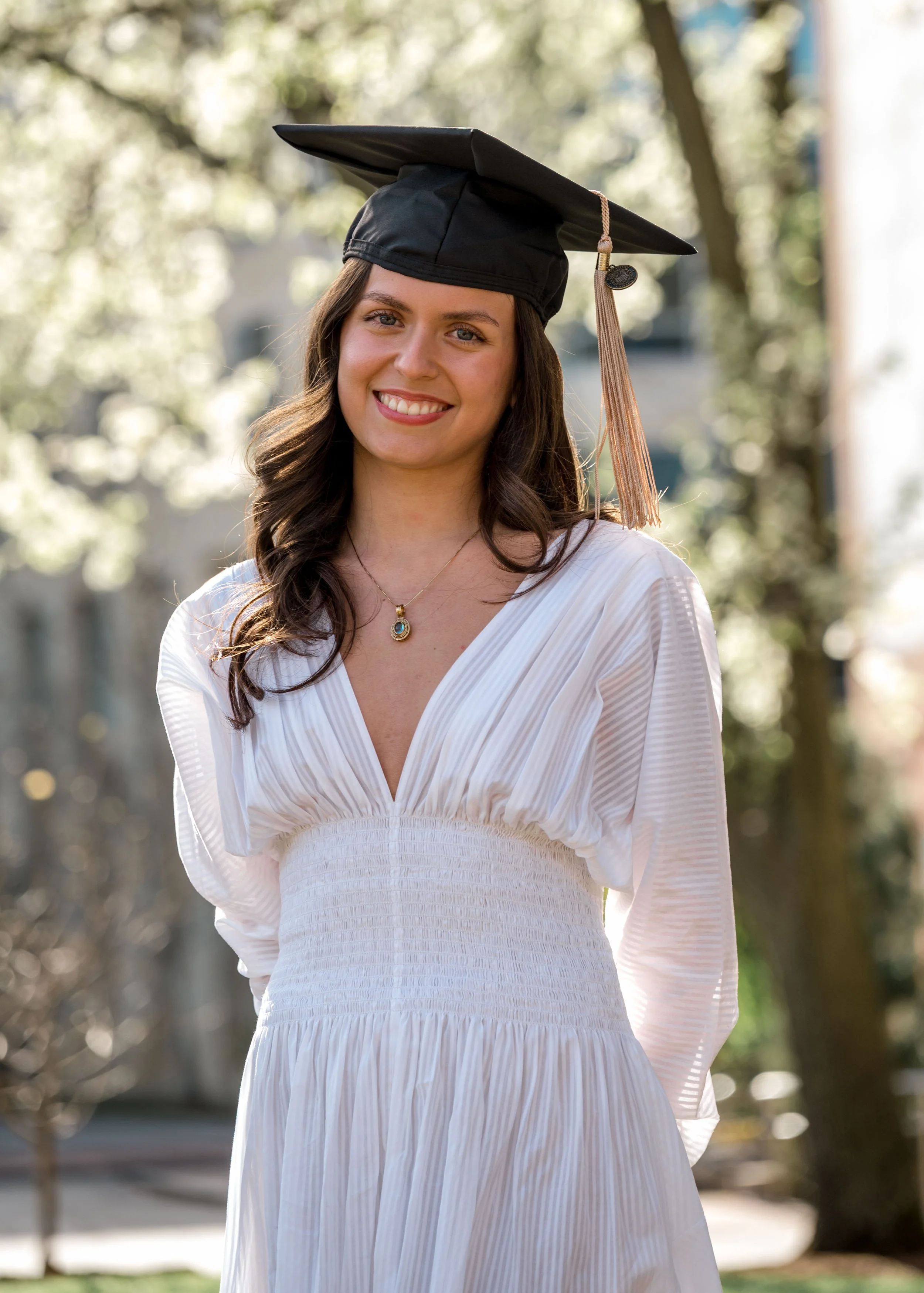 Young woman smiling outdoors wearing a white dress and a black graduation cap with a tassel.