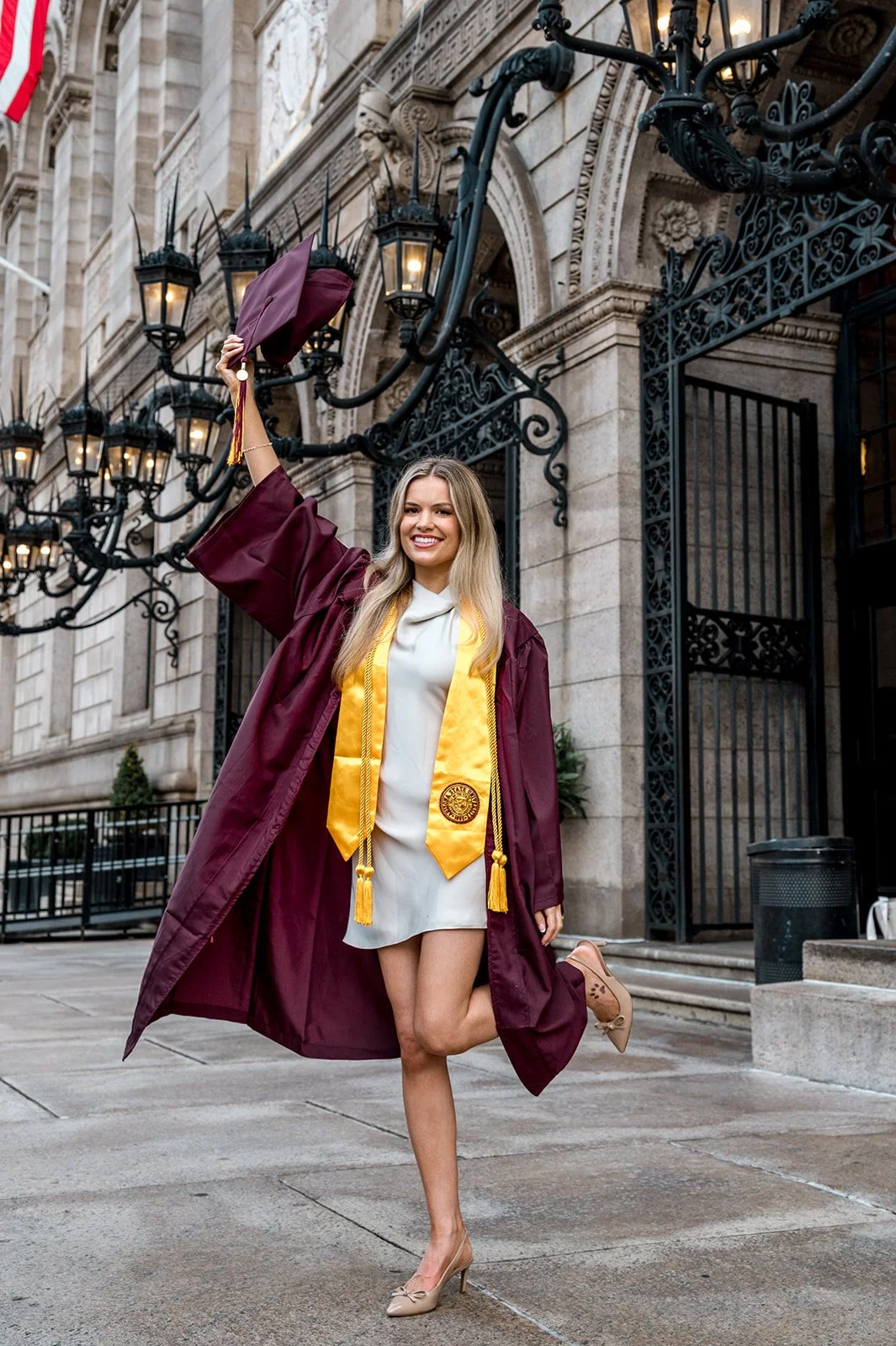 A young woman in a graduation cap and gown is smiling and celebrating outside a historic building, holding her cap in the air.