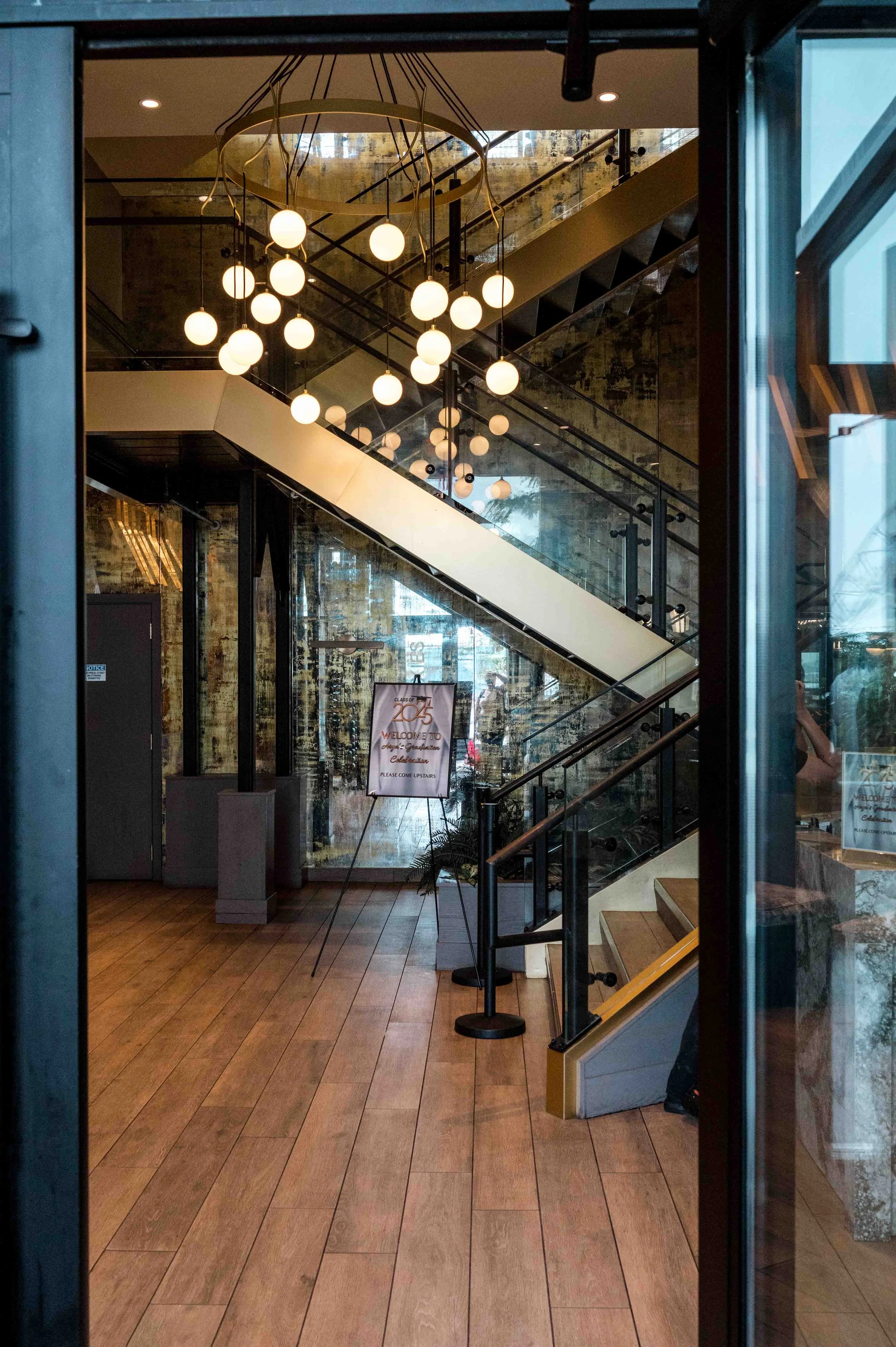 Interior view of a modern building lobby with a wooden floor, glass staircase, and a chandelier with multiple round lights hanging from the ceiling. There is a sign on a stand in the background and a large window reflecting the outside.