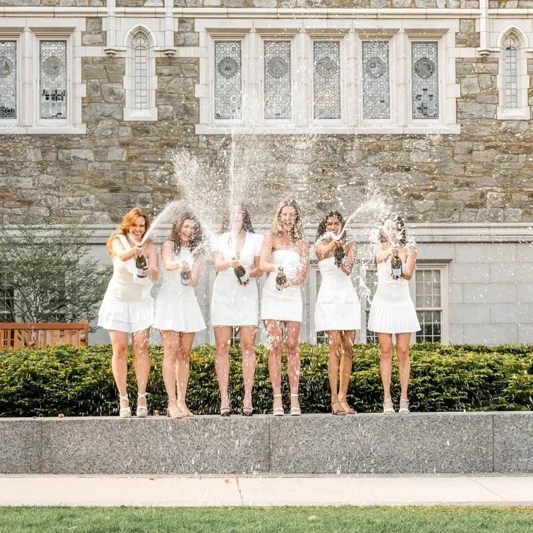 Six women in white dresses celebrating with champagne bottles on a fountain in front of a stone building.