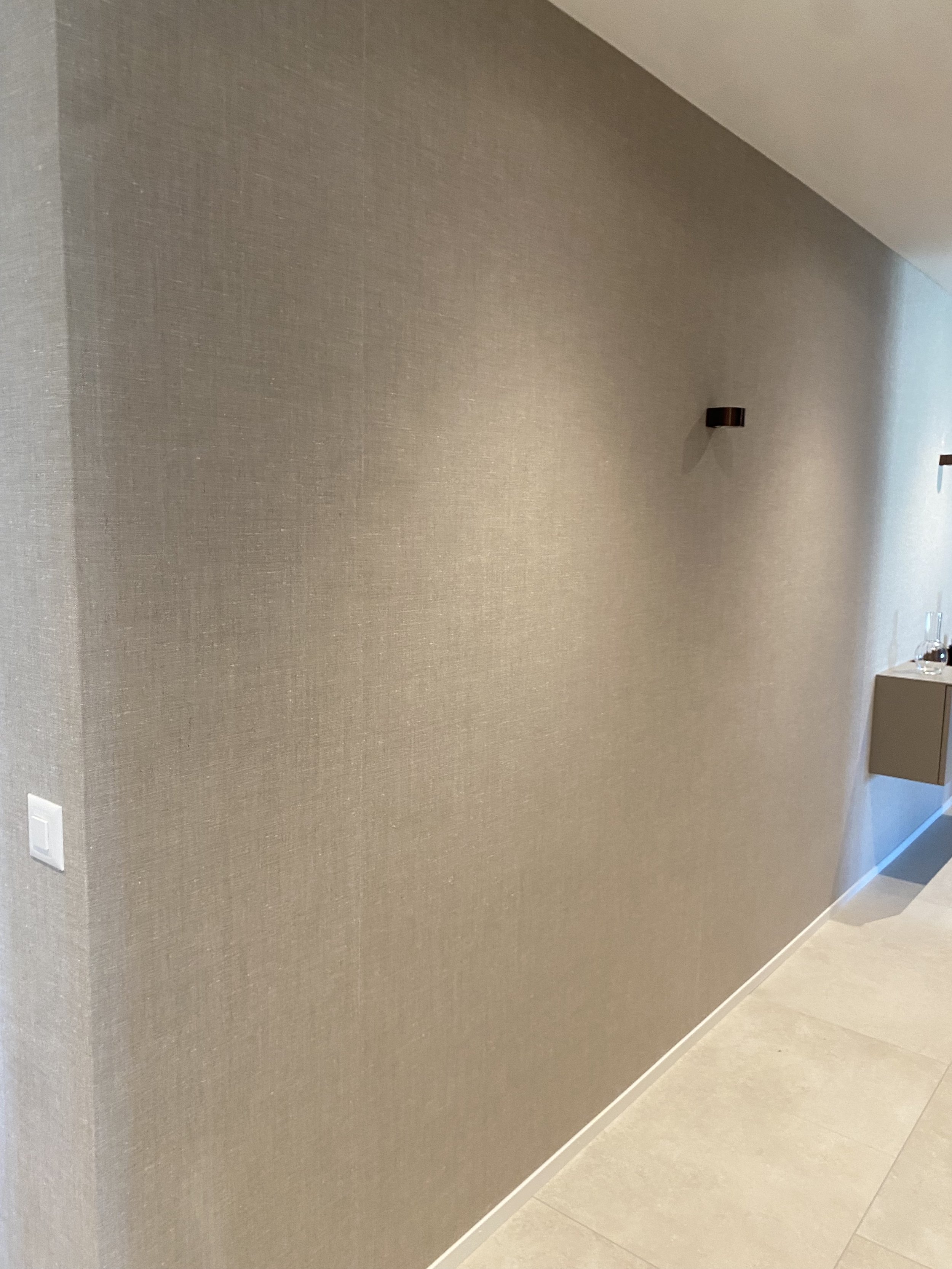 Ein grauer, texturierter Wandabschnitt in einem modernen Raum mit schwarzem Wandleuchter und einem kleinen beige farbigen Wandregal mit Dekorationsartikeln.