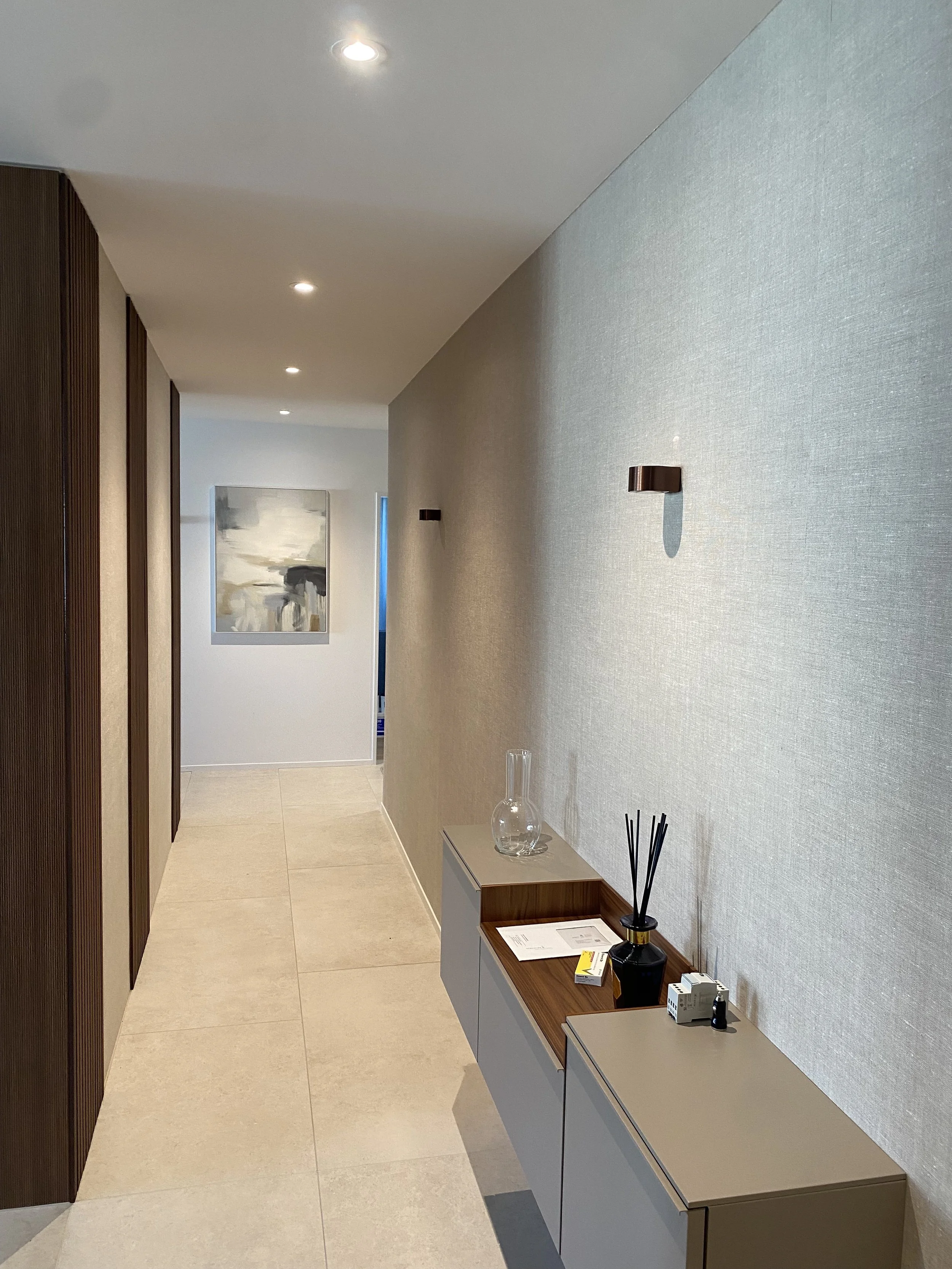 Moderner Flur mit weißem Boden, grauen Wänden, kleinen Wandleuchten und einem Gemälde an der Wand. Es gibt eine kleine Anrichte mit Dekorationsgegenständen wie eine Glasvase und einem Räucherstäbchenhalter.
