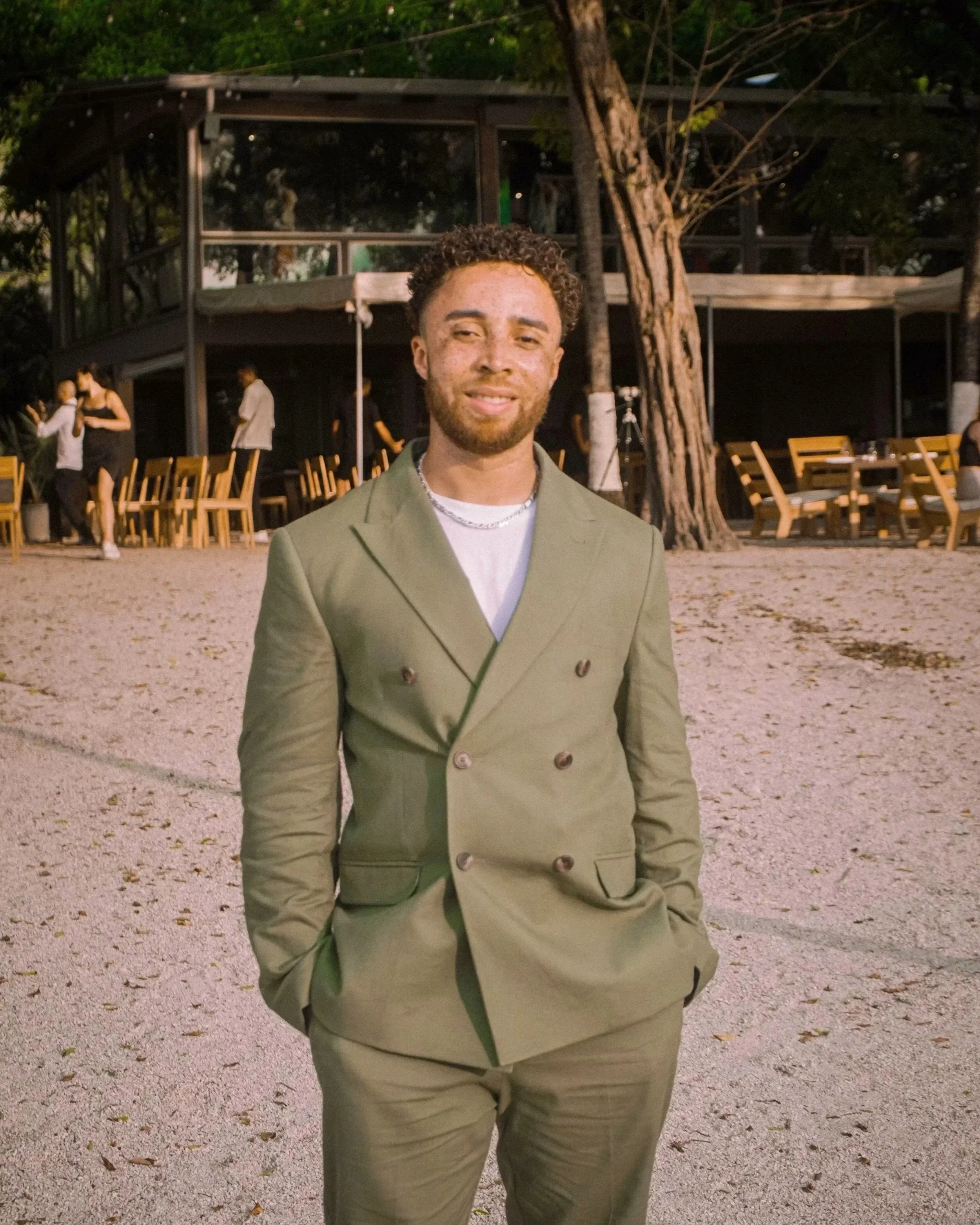 A young man with curly hair and a beard standing outdoors at dusk, wearing a green double-breasted suit and smiling at the camera.