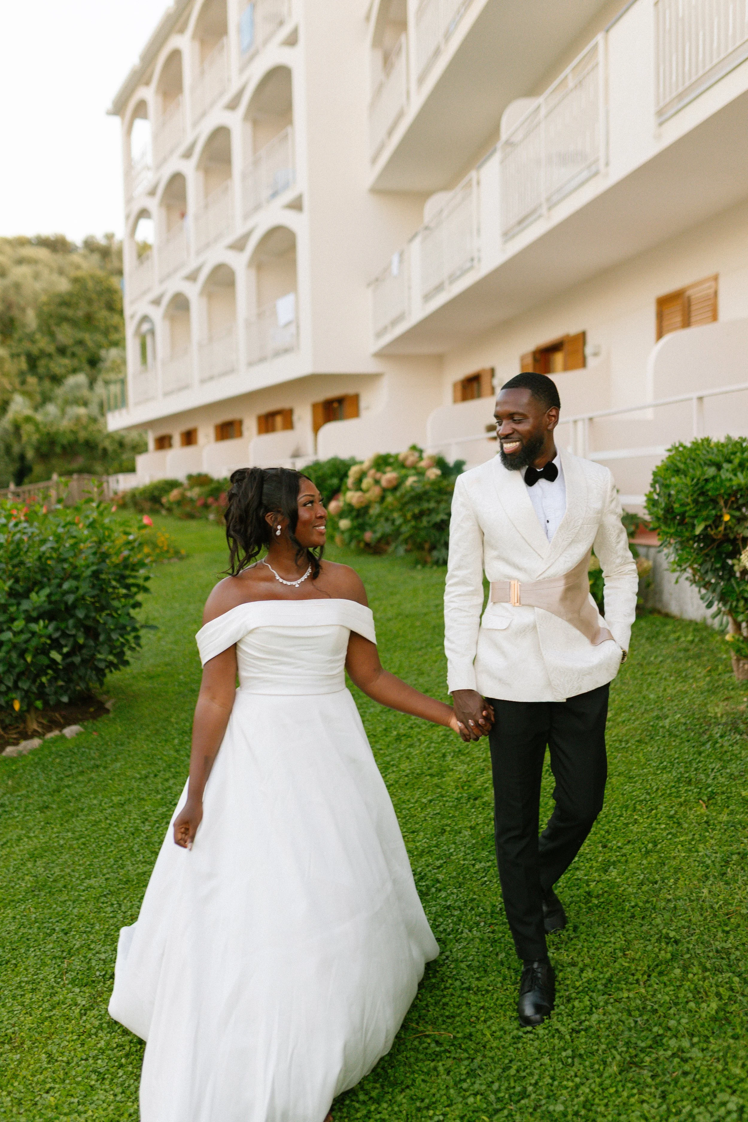 A newly married couple walking hand-in-hand outside a white building with multiple balconies, during daytime.