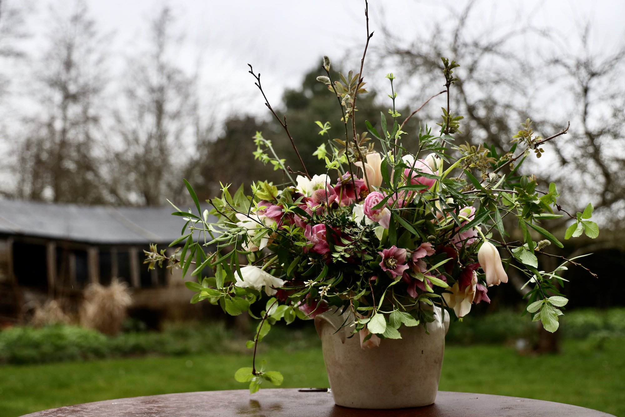 Flower arrangement in a pot with pink and white blossoms, greenery on outdoor table, garden background.