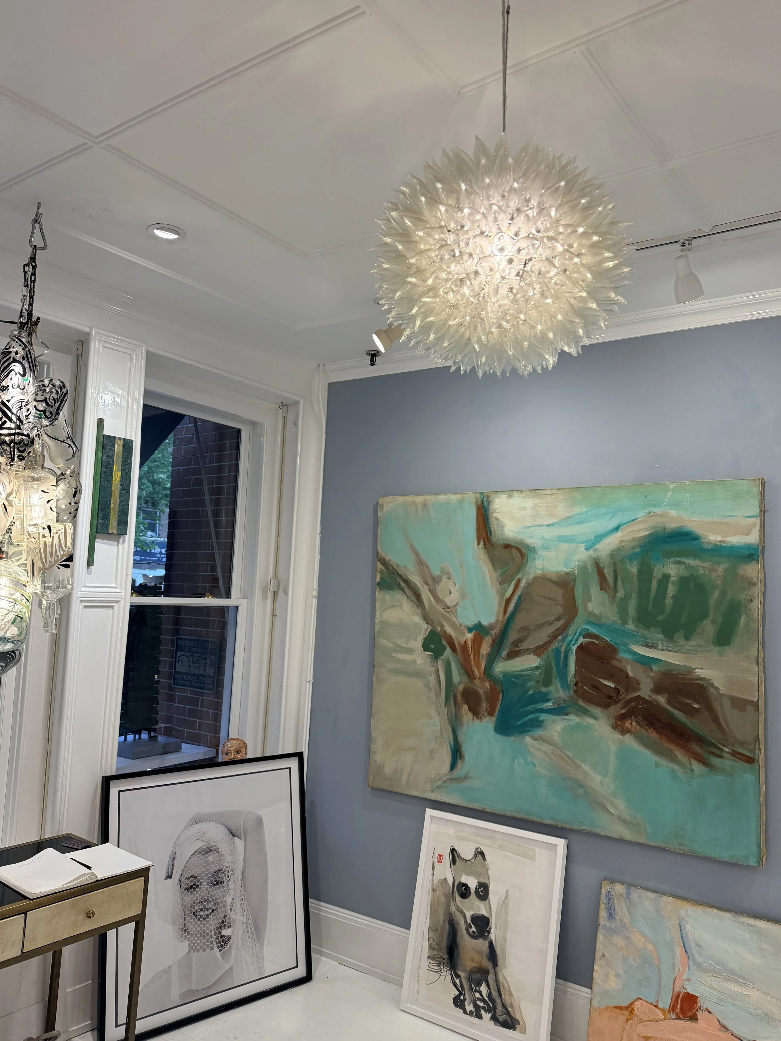 View of an art gallery corner with various paintings leaning against the wall, including an abstract piece with blue, beige, and orange colors, a portrait of a dog, and a black-and-white framed photo of a woman with a veil.