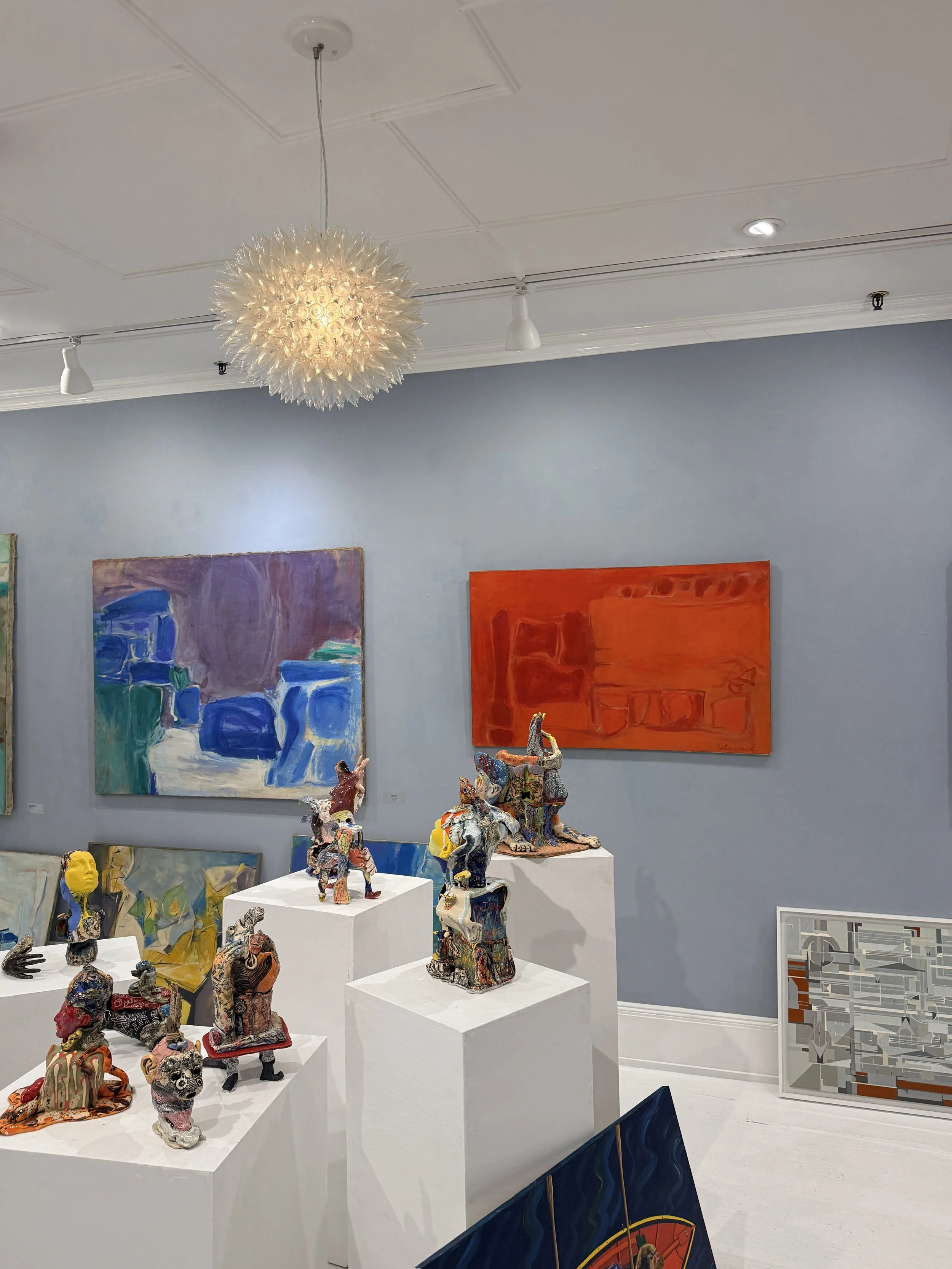Art gallery with colorful abstract paintings on the walls and ceramic sculptures displayed on white pedestals in the center of the room. A decorative ceiling light fixture hangs from the ceiling.