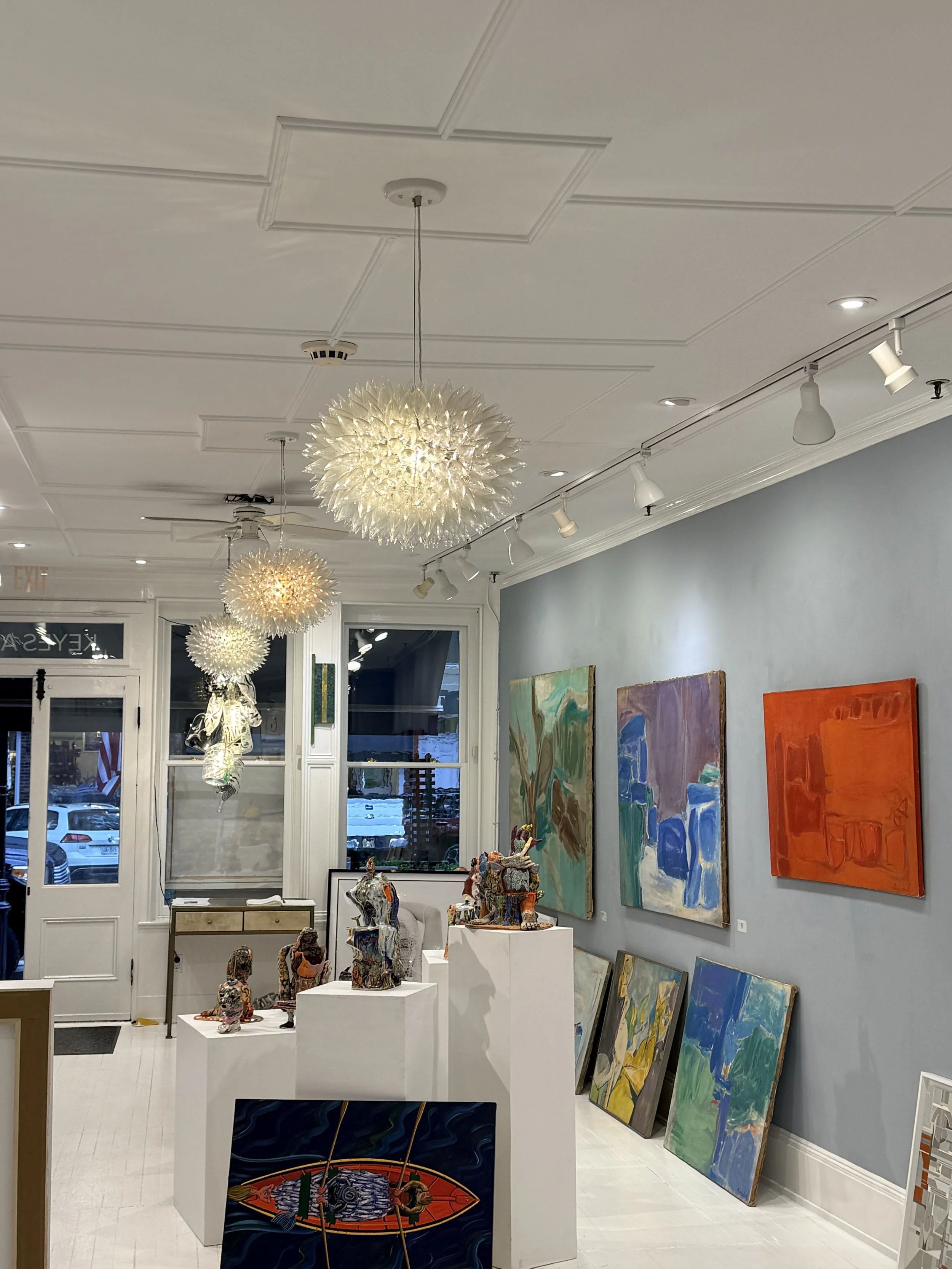 An art gallery with colorful paintings on the wall and sculptures on white pedestals, illuminated by ceiling lights and hanging chandeliers.