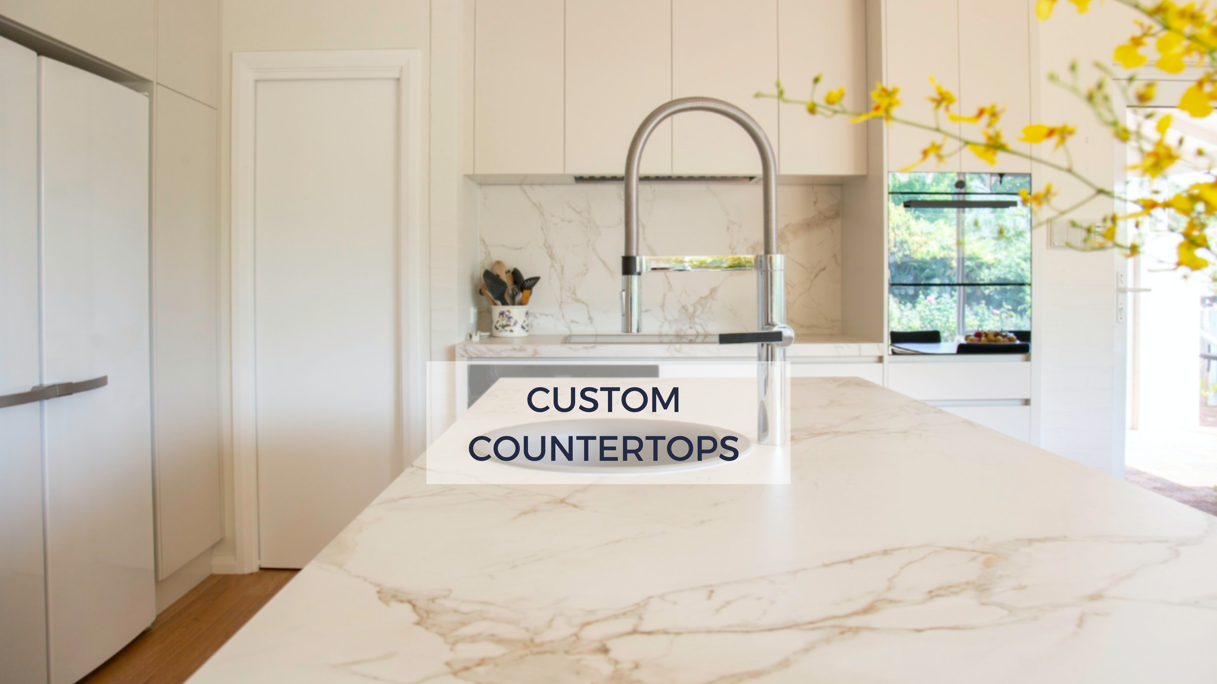 Modern kitchen with white marble countertops, a sink with a high-arc faucet, and a window overlooking a garden. Text overlay says "Custom Countertops."