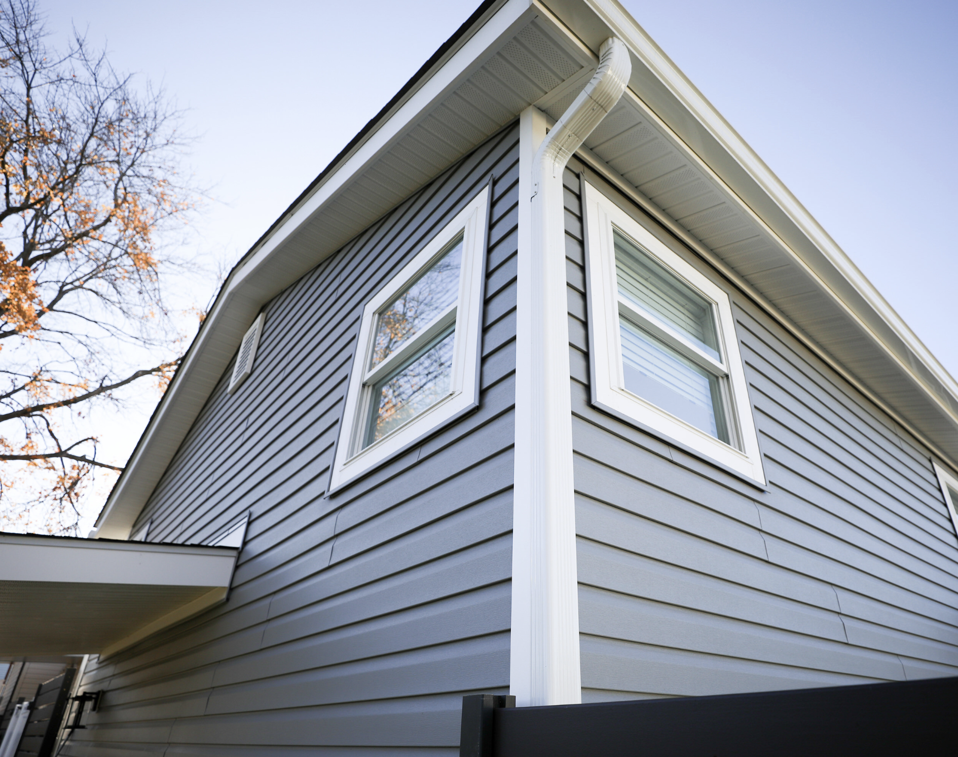 Close-up of the corner of a modern house with blue siding, white trim, and double-pane windows, during daytime with a clear sky in the background.