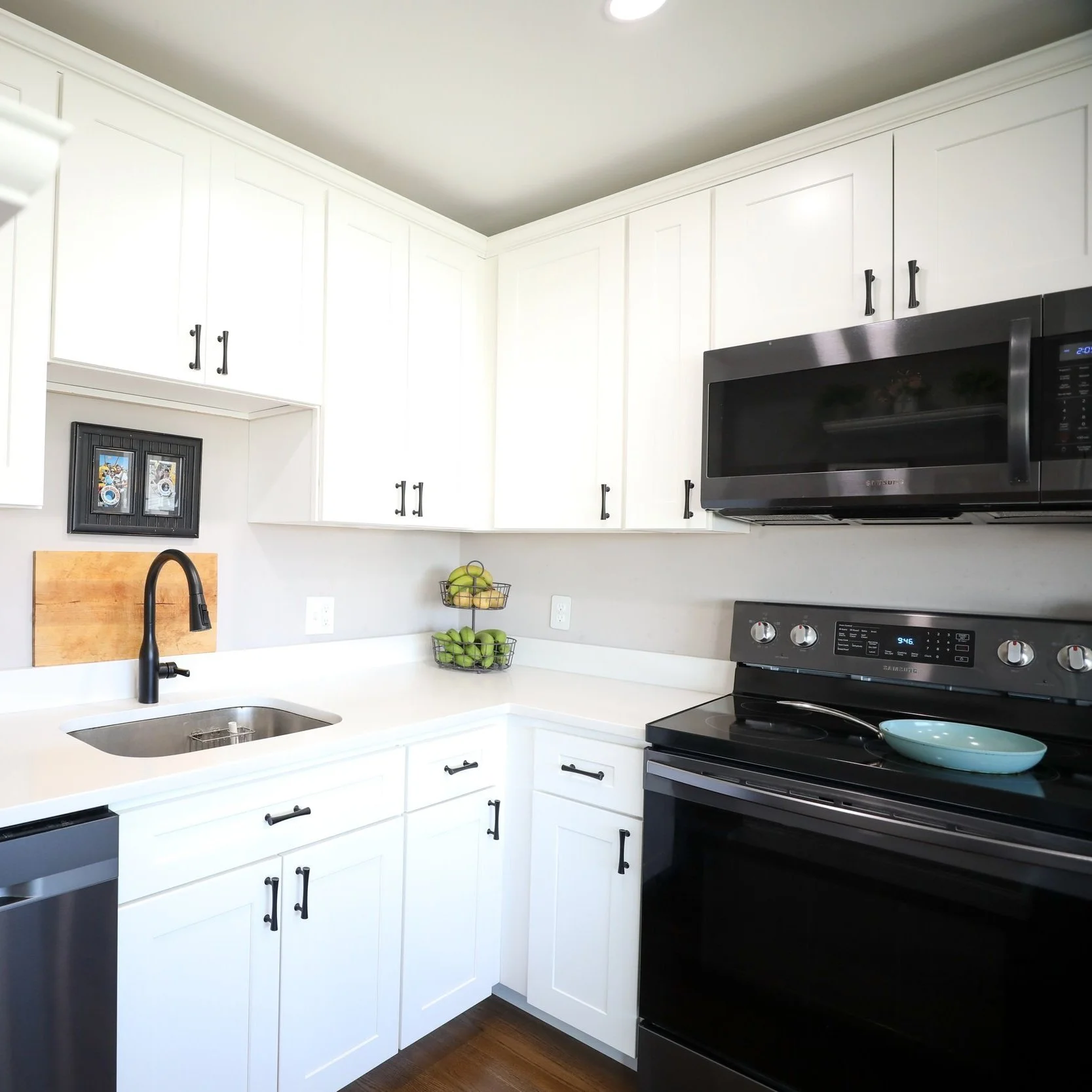 White kitchen with black cabinet handles, black microwave, black stove, and a wire fruit basket on the counter.