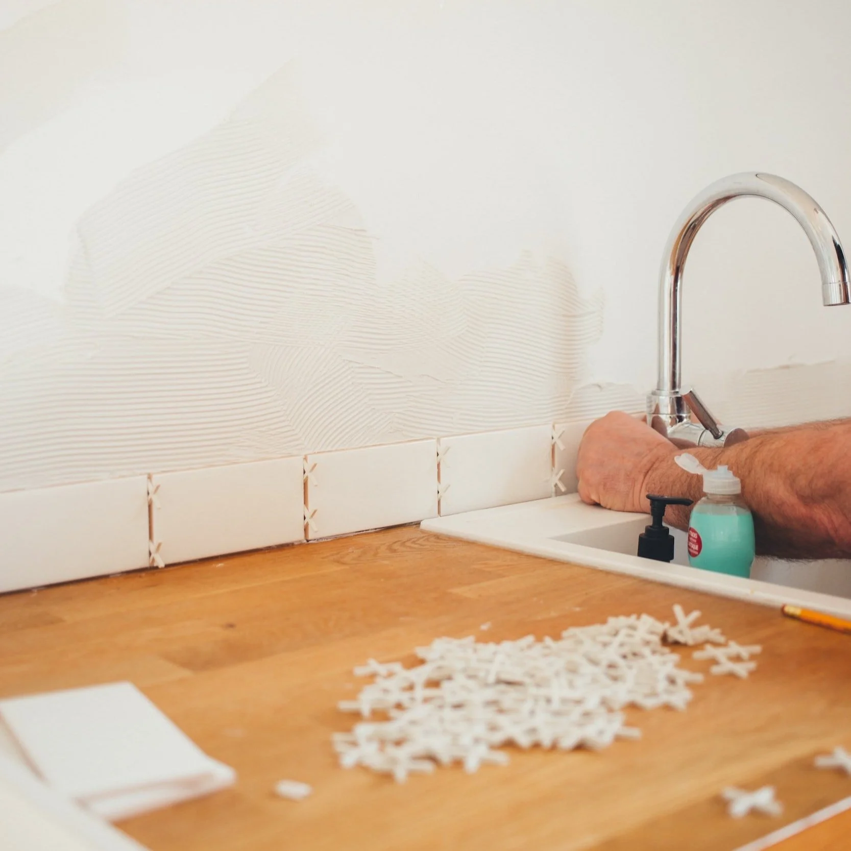 A person installing white ceramic tiles at a kitchen sink, with a pile of tile spacers on the wooden countertop.