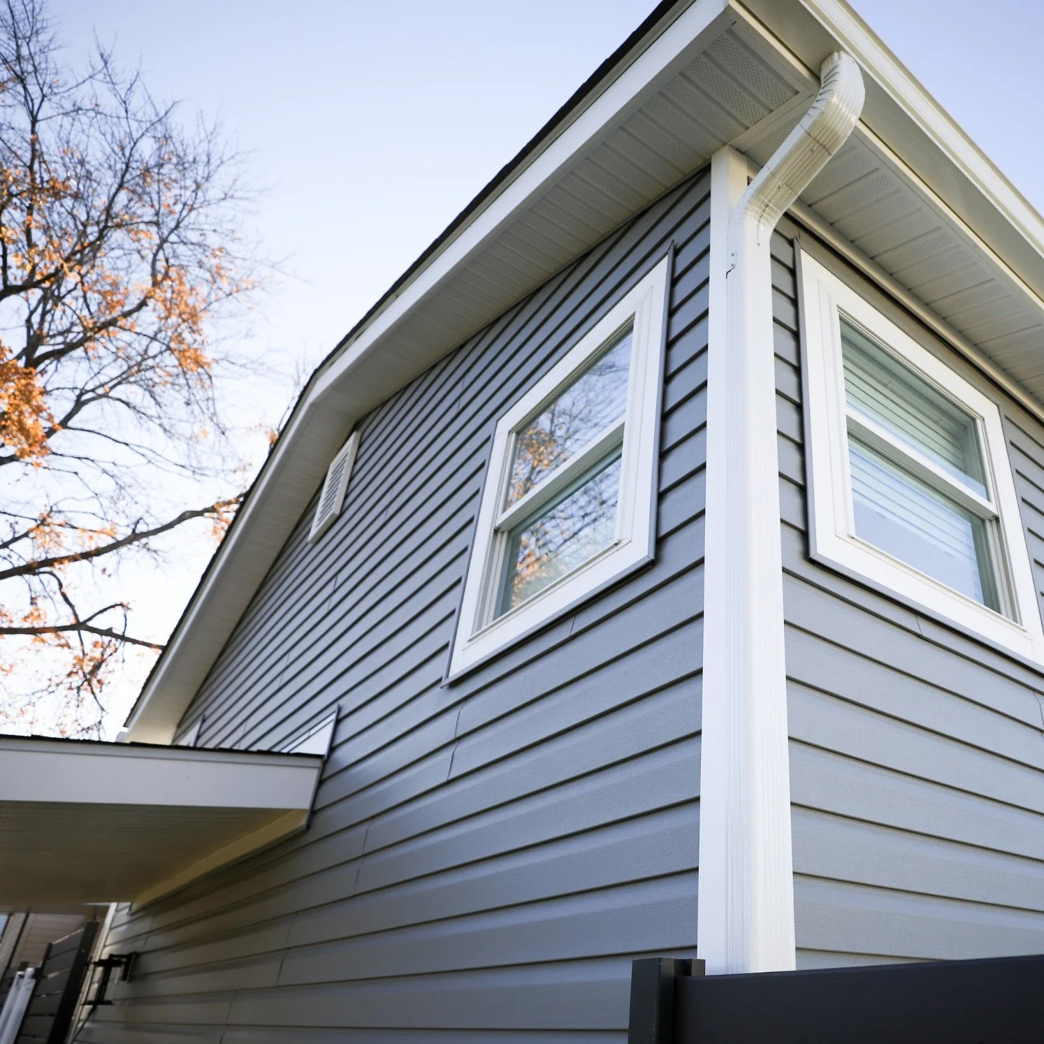 Close-up of a modern house exterior with gray siding, white window frames, and a white gutter pipe, with trees visible in the background.