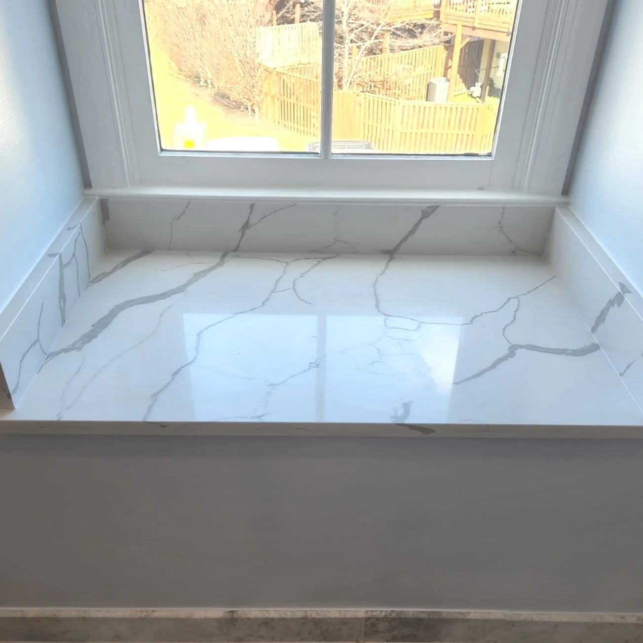 Close-up of a marble window sill with white and gray veining, near a large window with a view of a backyard fence.