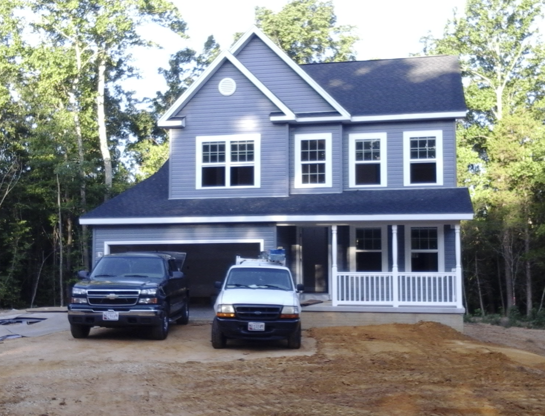 New two-story gray house with white trim, front porch, and several windows, with two vehicles parked in front, surrounded by trees and a dirt driveway.