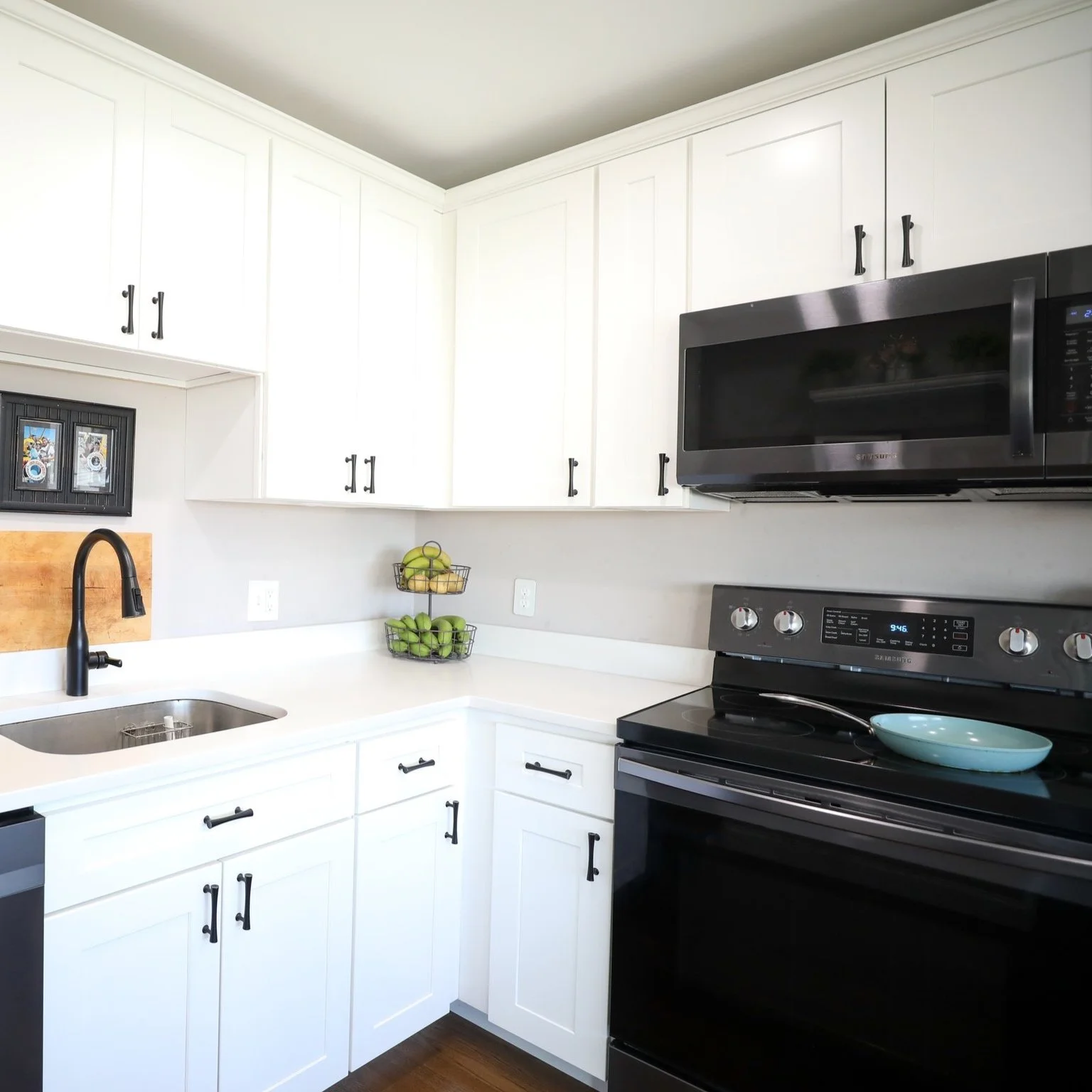 Modern kitchen with white cabinets, black hardware, black stove, black microwave, and a sink with a curved black faucet. Green apples in a wire basket on the counter.