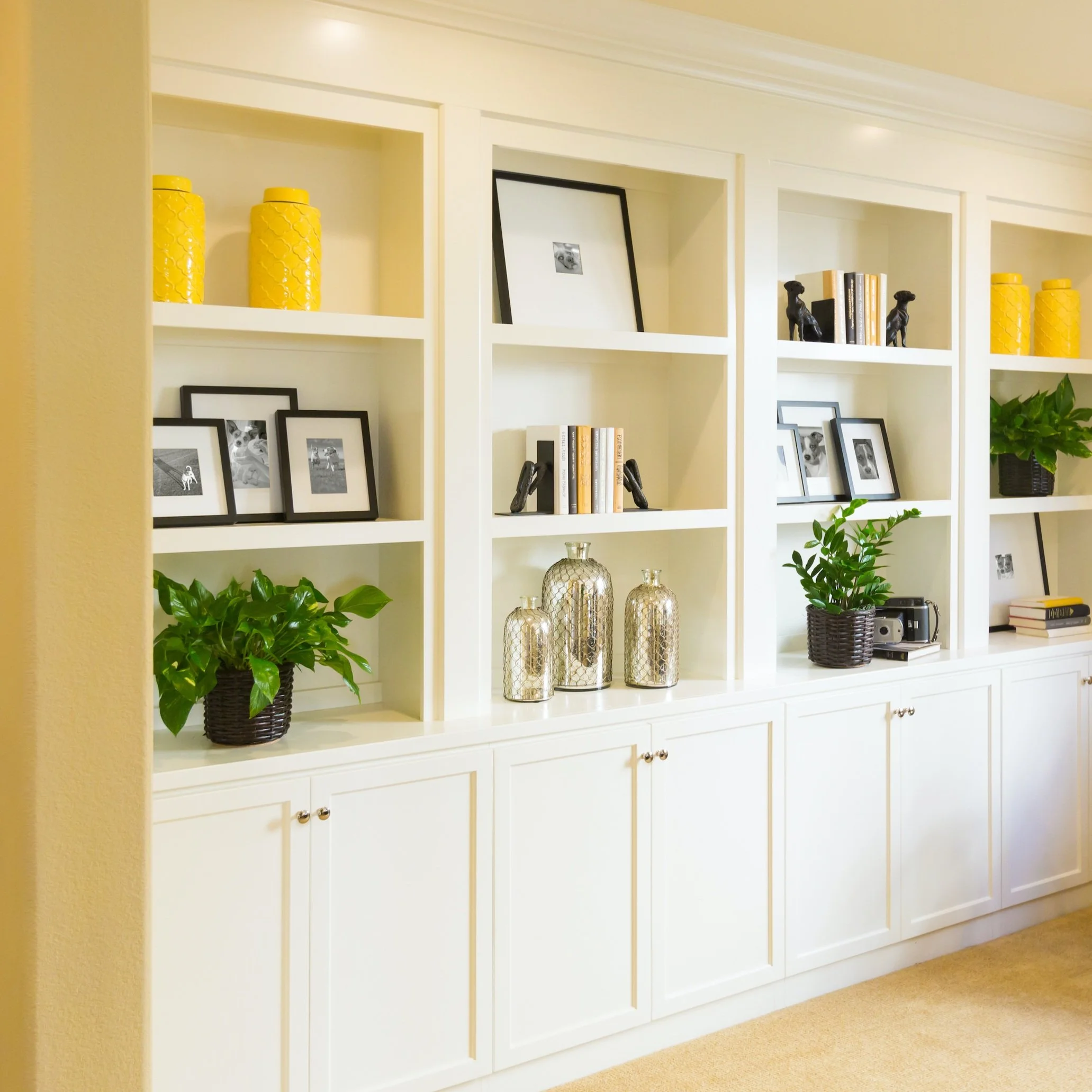 White built-in bookshelf with framed photos, black and white picture frames, yellow vases, potted plants, books, decorative silver bottles, and a vintage camera.