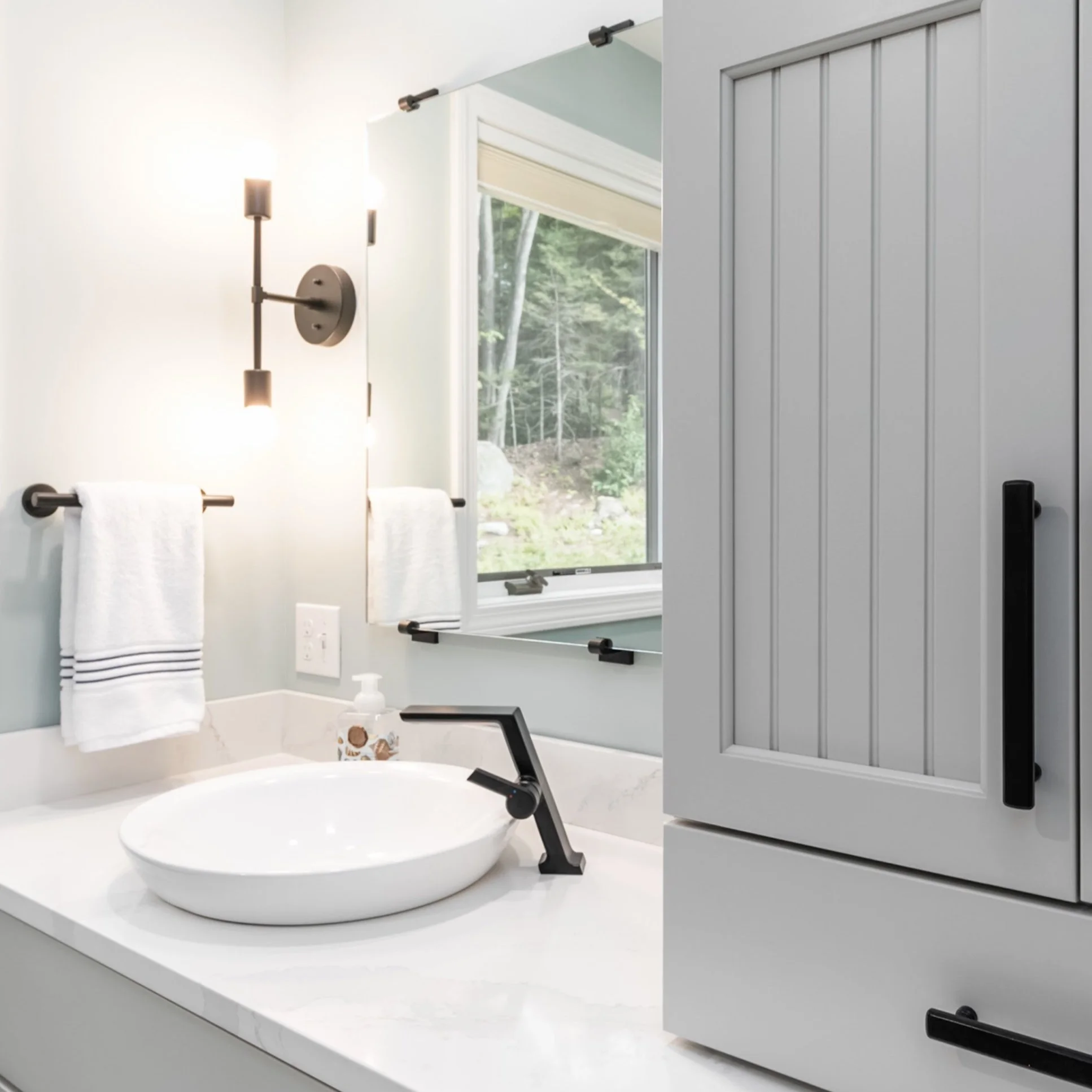 Modern bathroom with white countertop, oval sink, black faucet, wall-mounted light fixture, towel bar with white towels, window with view of trees, and gray cabinetry.