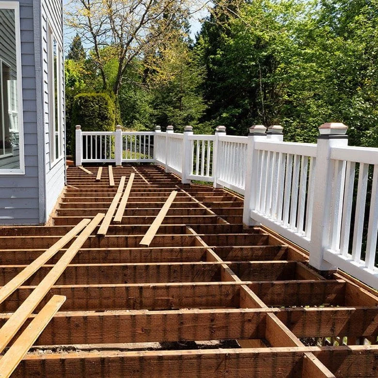 Outdoor deck under construction with wooden framing and a white railing, next to a house with siding, surrounded by green trees.