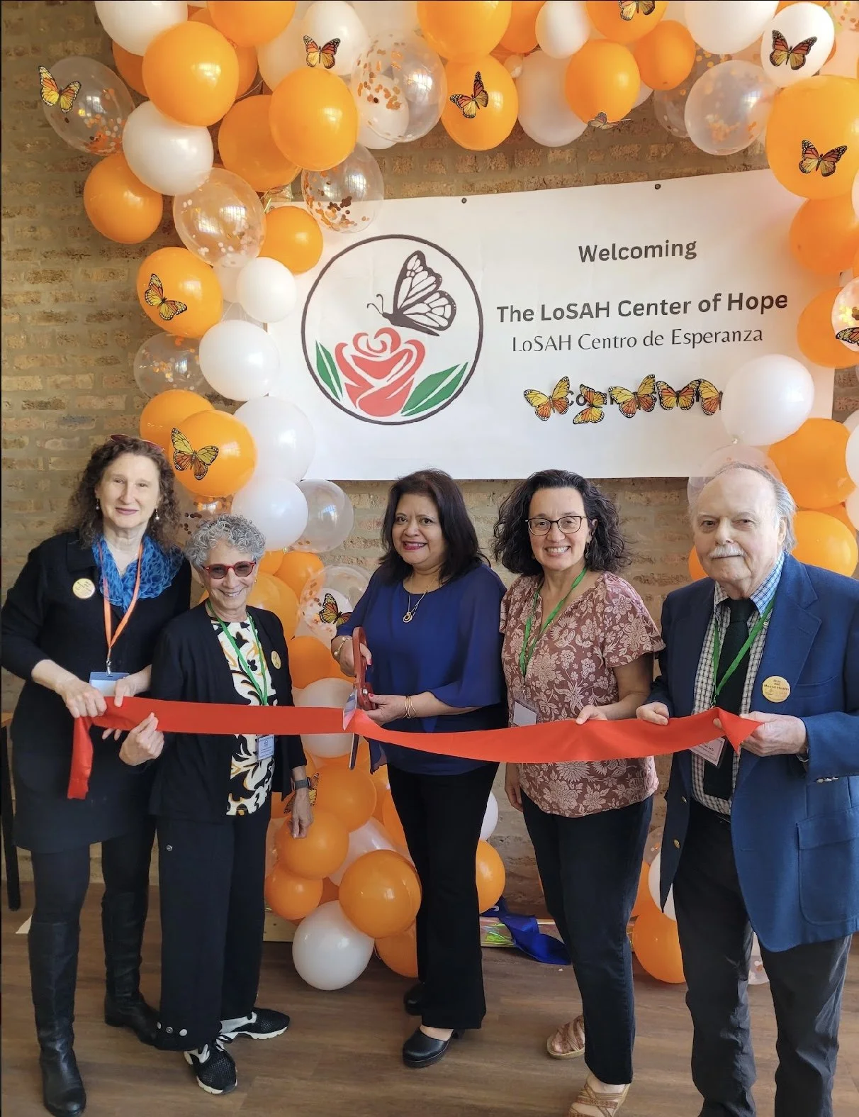 Five people participating in a ribbon-cutting ceremony in front of a decorated wall with a sign that reads "Welcoming The LoSAH Center of Hope" in English and Spanish, with orange and white balloons and butterfly decorations.