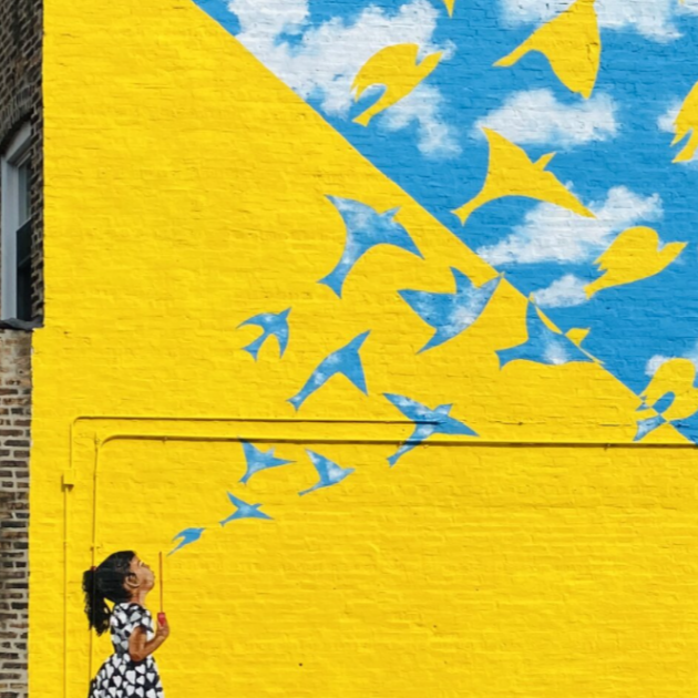 A girl in a black and white dress releases blue and yellow painted paper birds on a yellow and blue mural wall.